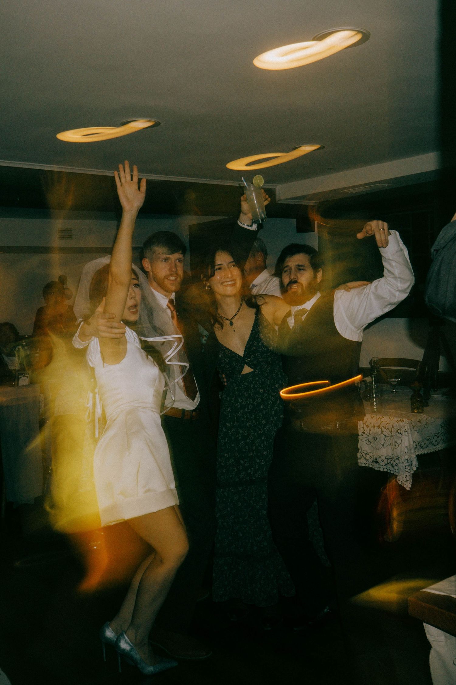 Wedding guests dance enthusiastically under ambient ceiling lights at the reception.
