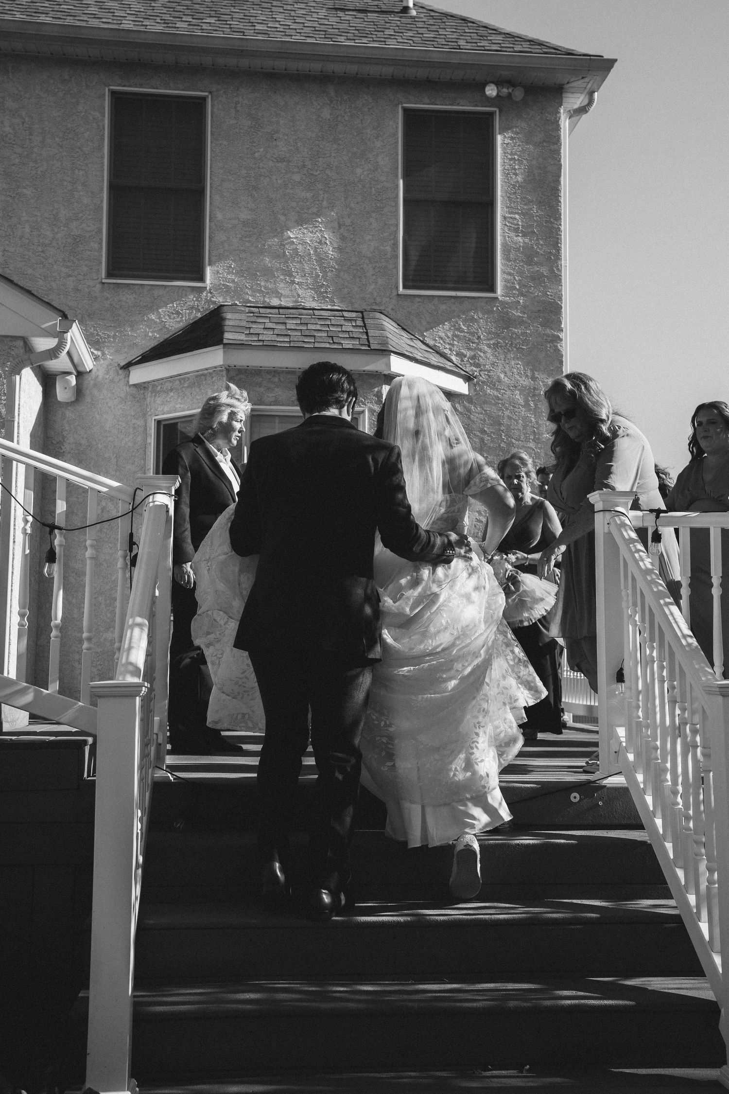 Wedding party ascends white stairs outside brick building in black and white photograph.