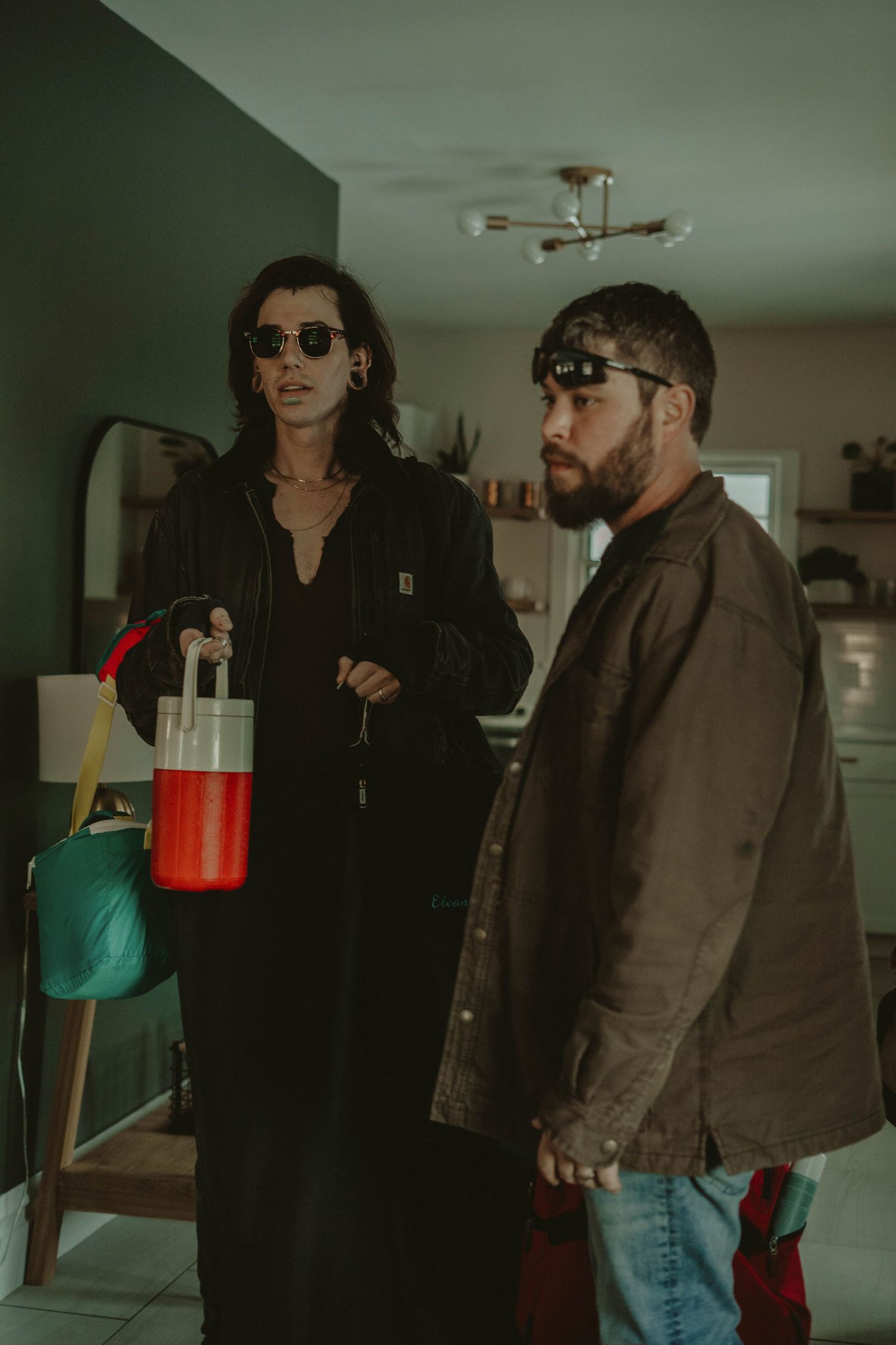 Two people in dark clothing and sunglasses stand indoors holding beverages.
