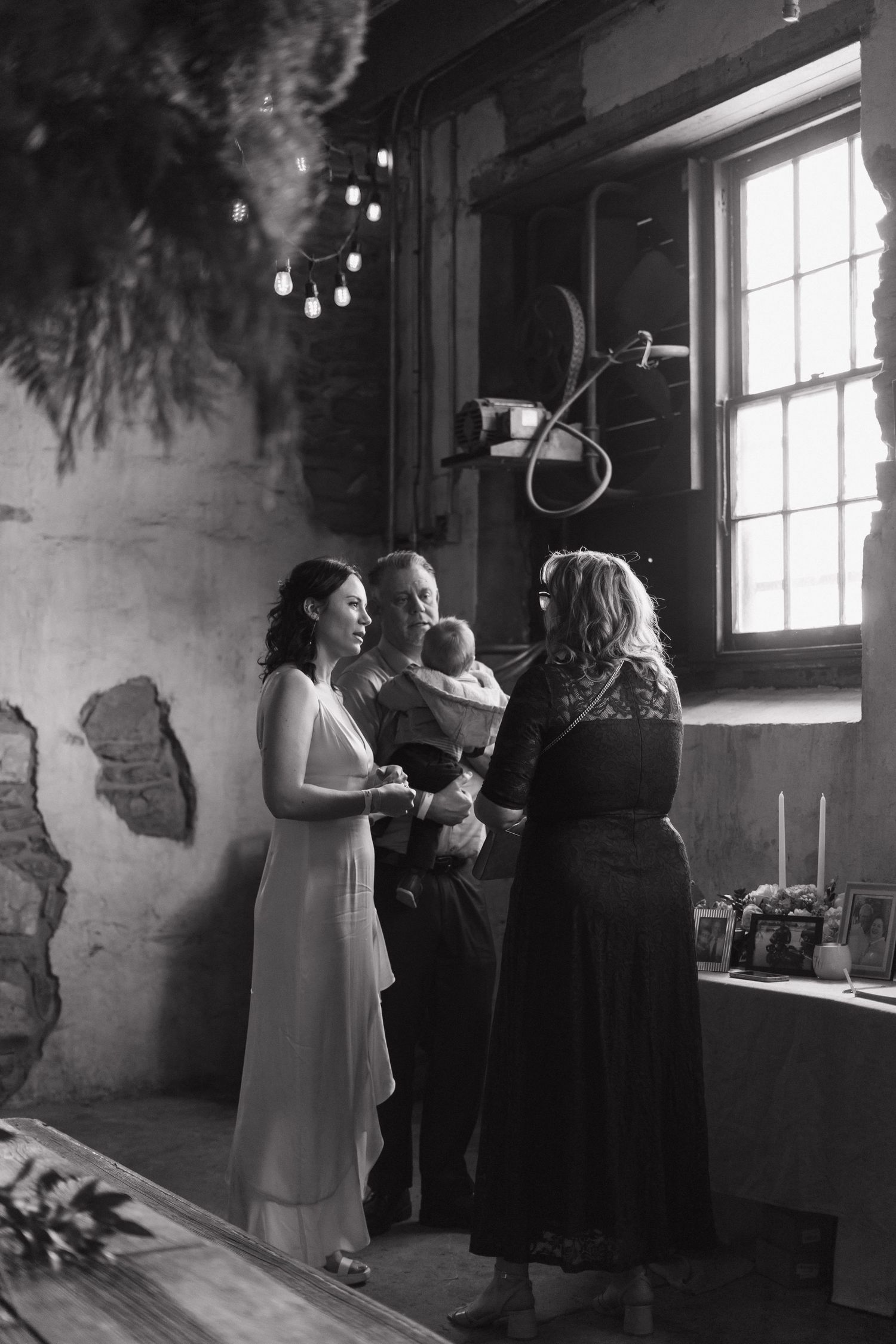 A black and white wedding ceremony photo showing a small intimate gathering by a sunlit window in a rustic interior setting.