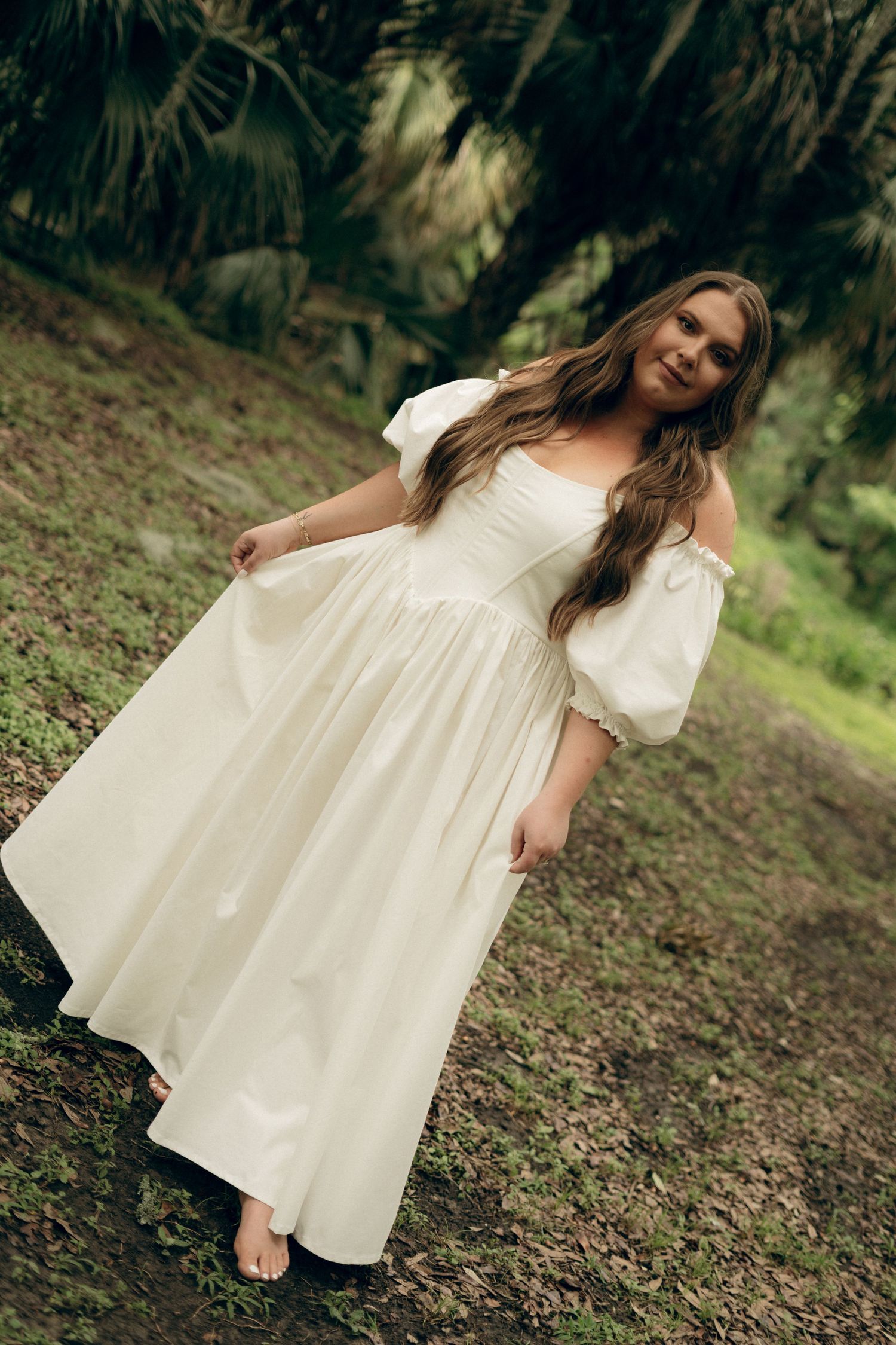 A person in a flowing white dress poses gracefully on a grassy hillside in a natural wooded setting.