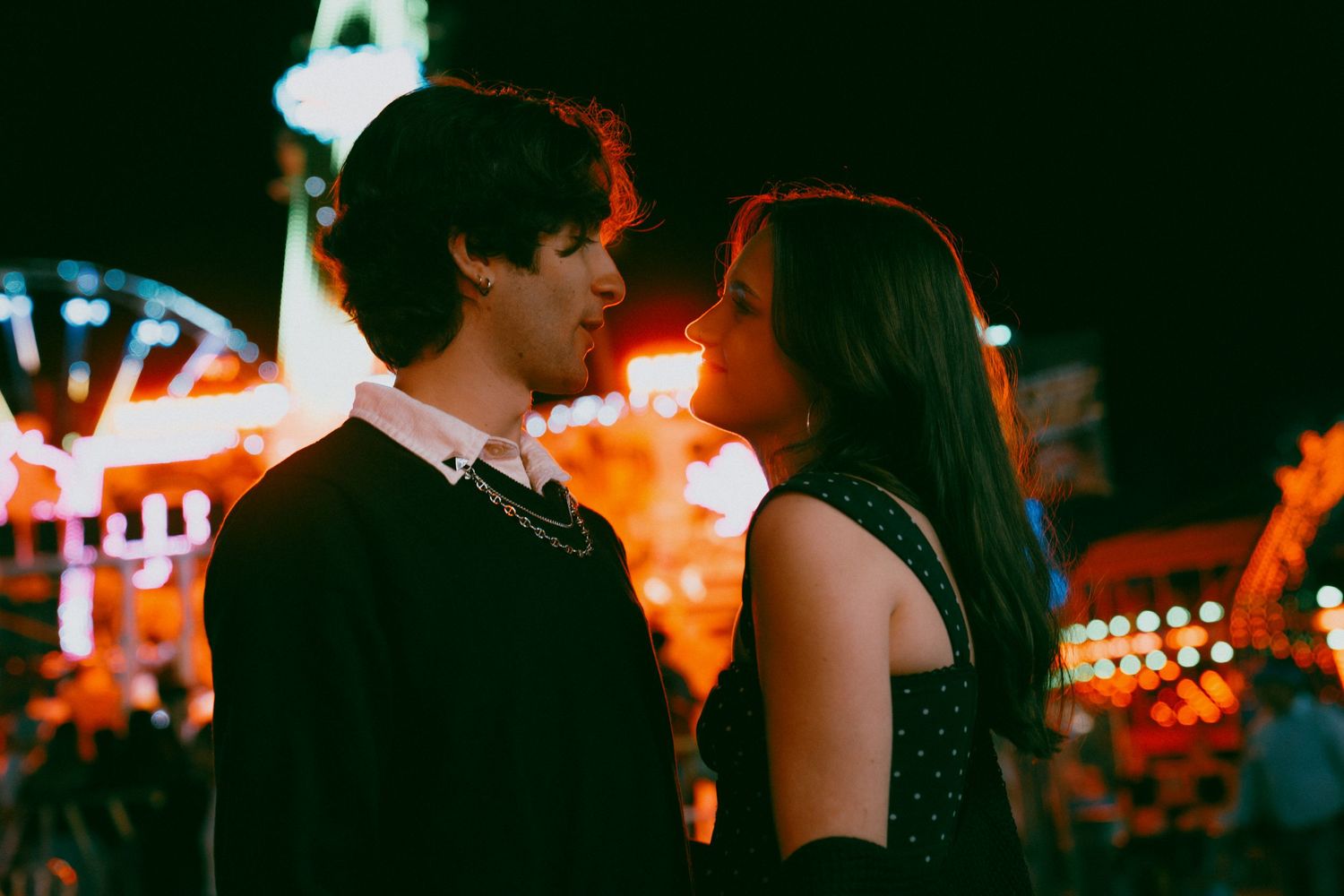 A romantic couple stands close together at night with colorful carnival lights glowing in the background.