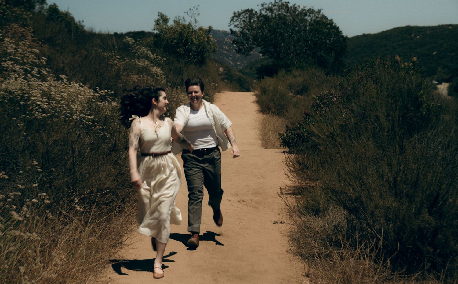 A couple walks together on a dusty hiking trail surrounded by green brush and shrubs on a sunny day.