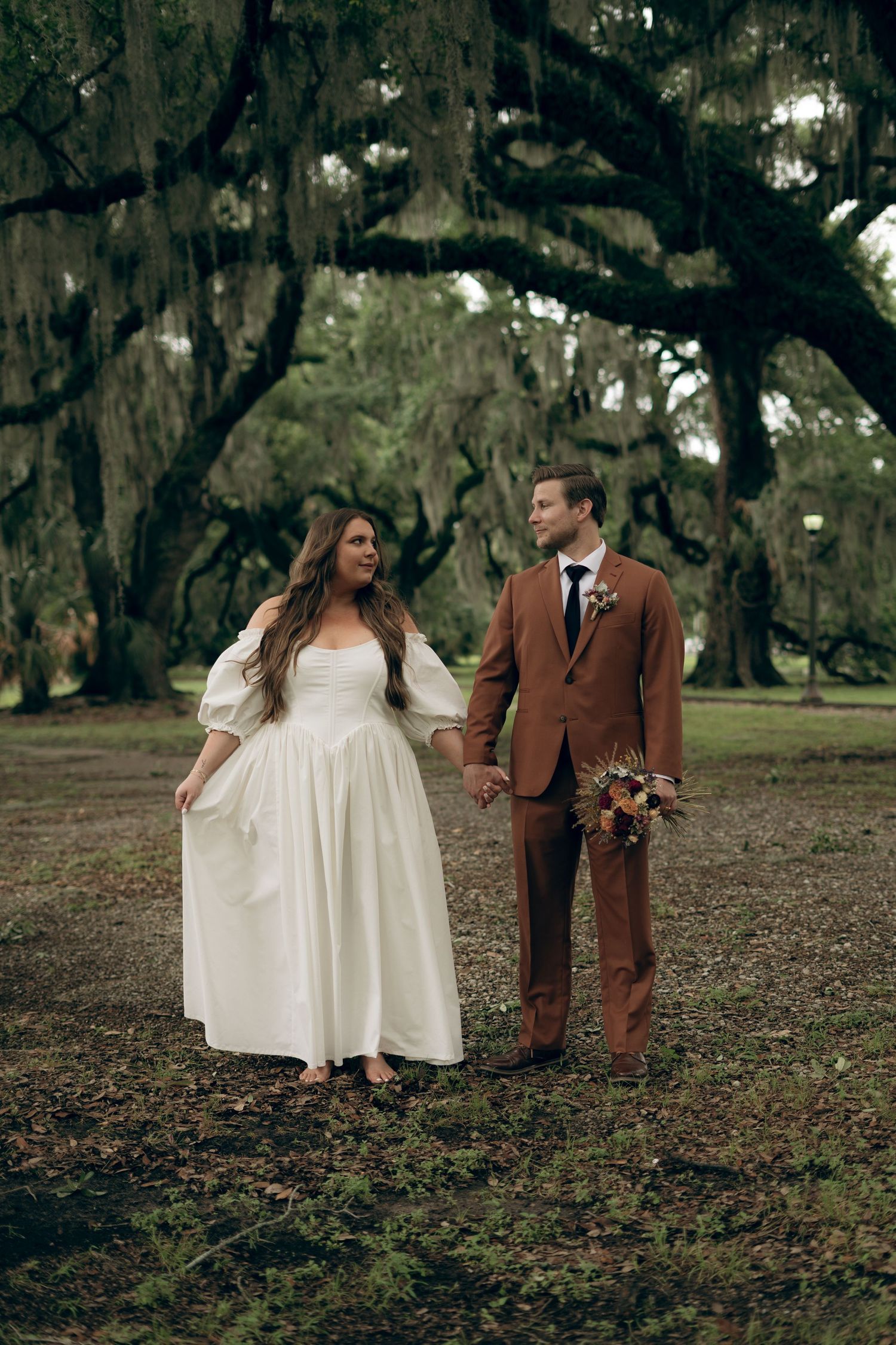 A couple in wedding attire poses among moss-draped oak trees in a dreamy outdoor setting.