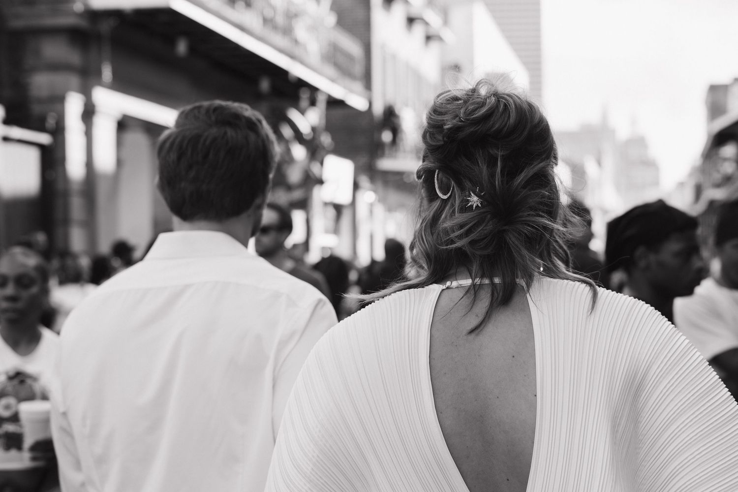 Black and white photo showing a person's back with a deep V-cut white dress in a crowded street scene.