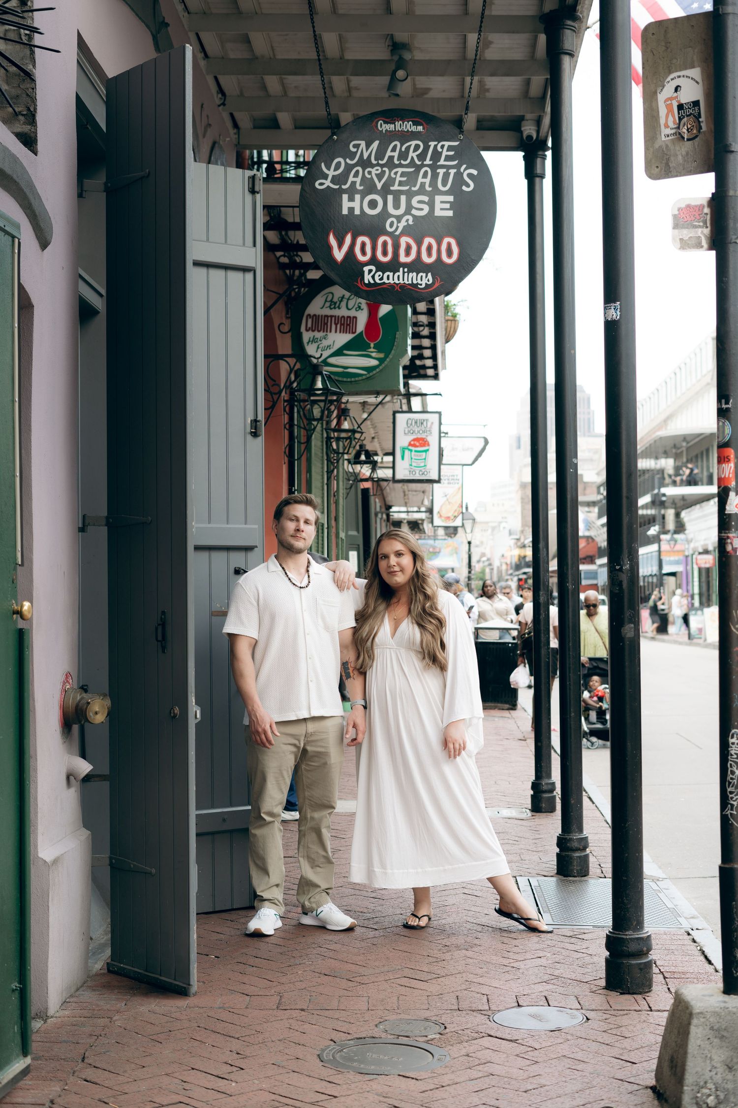 People walk past historic buildings with green columns and black doors along a brick sidewalk on Bourbon Street in New Orleans.