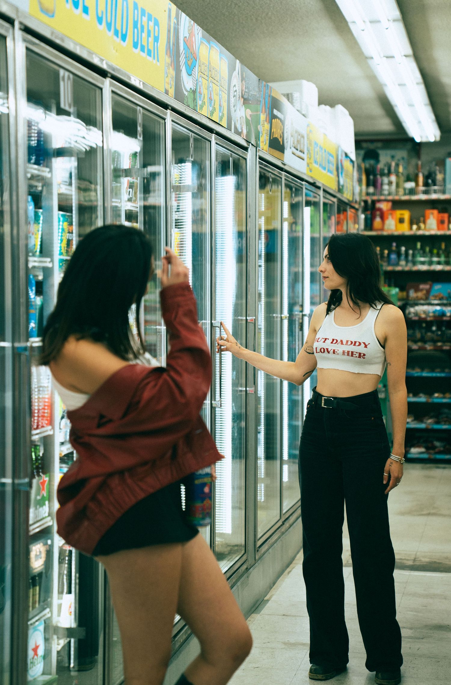 Two people browse refrigerated beverages in a convenience store with glass-door coolers and fluorescent lighting.