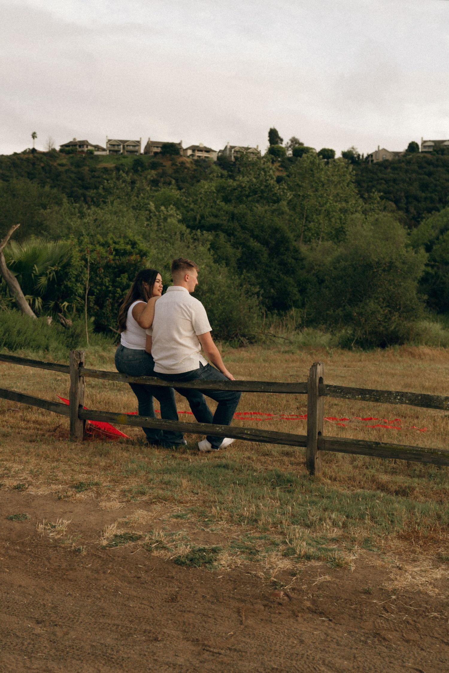 Couple sitting together on a rustic wooden fence overlooking a green hillside with homes in the background.