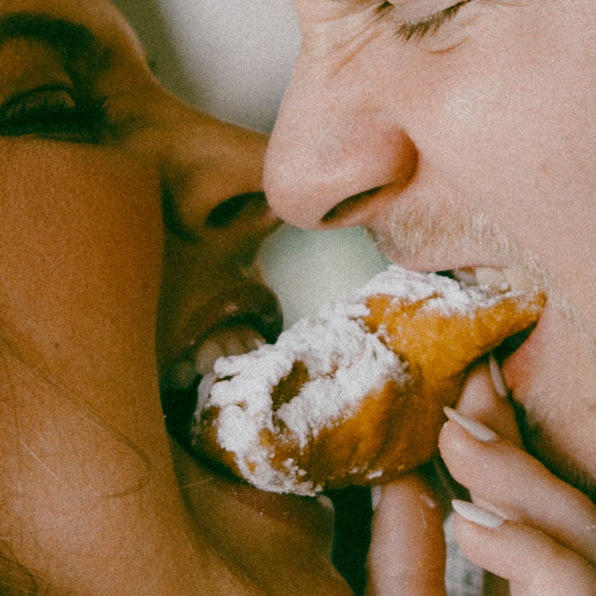 Two people sharing a powdered pastry or donut together in an intimate close-up shot.