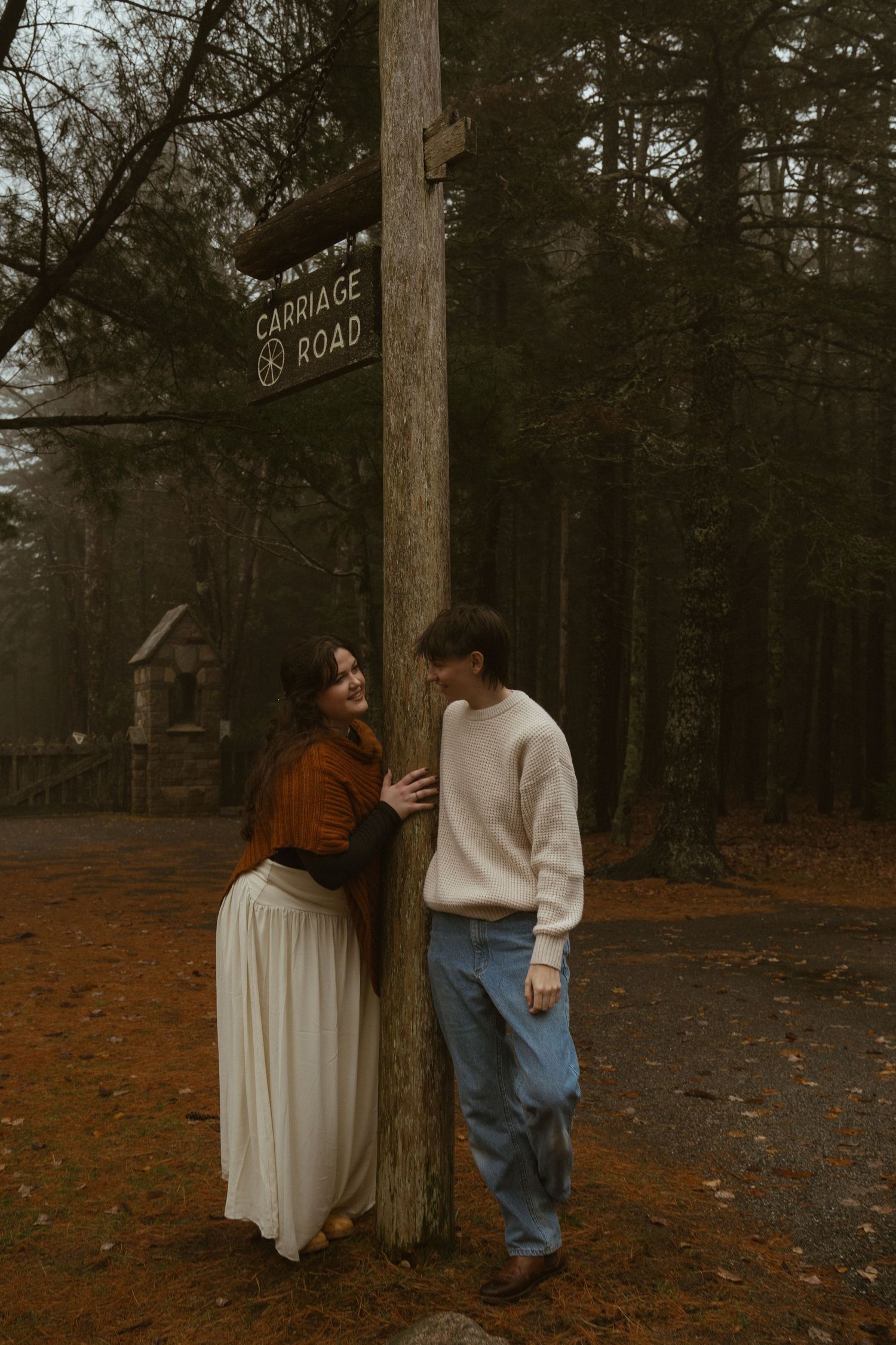 Couple sharing a romantic moment leaning against a wooden Carriage Road sign post in an autumn forest.