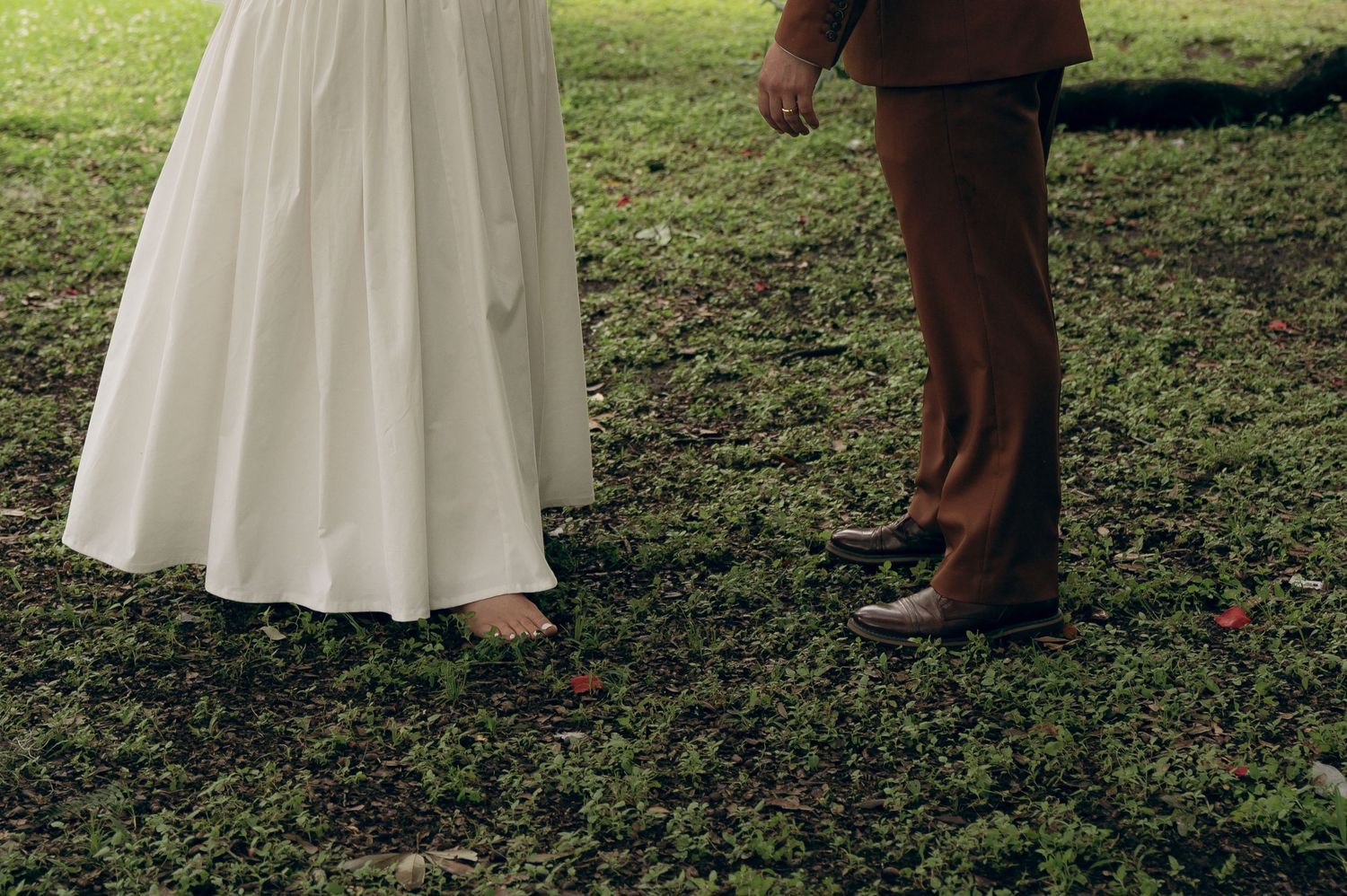 A long white formal dress and brown dress shoes stand on grass in a cropped wedding photography composition.