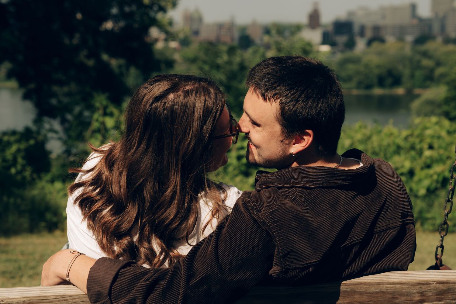 A couple in casual attire share an intimate moment while sitting outdoors with a scenic view behind them.
