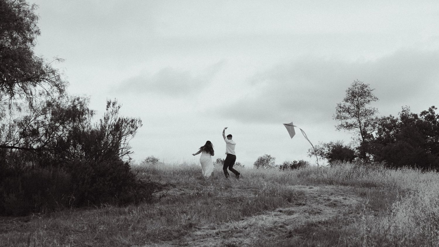 Two people joyfully running and flying a kite in an open field on a cloudy day in black and white.