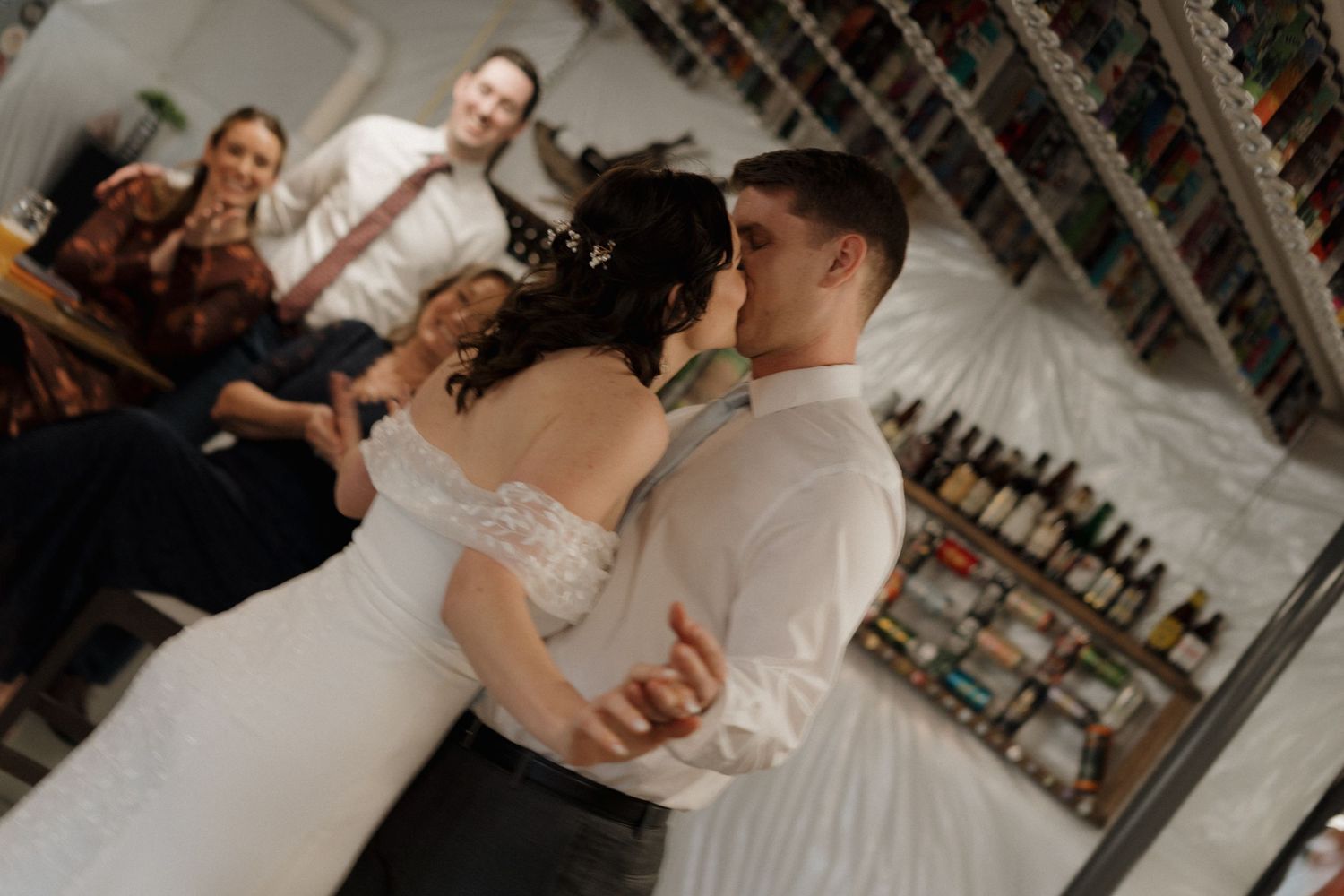 A couple shares their first dance at their wedding reception while guests watch and smile from behind.