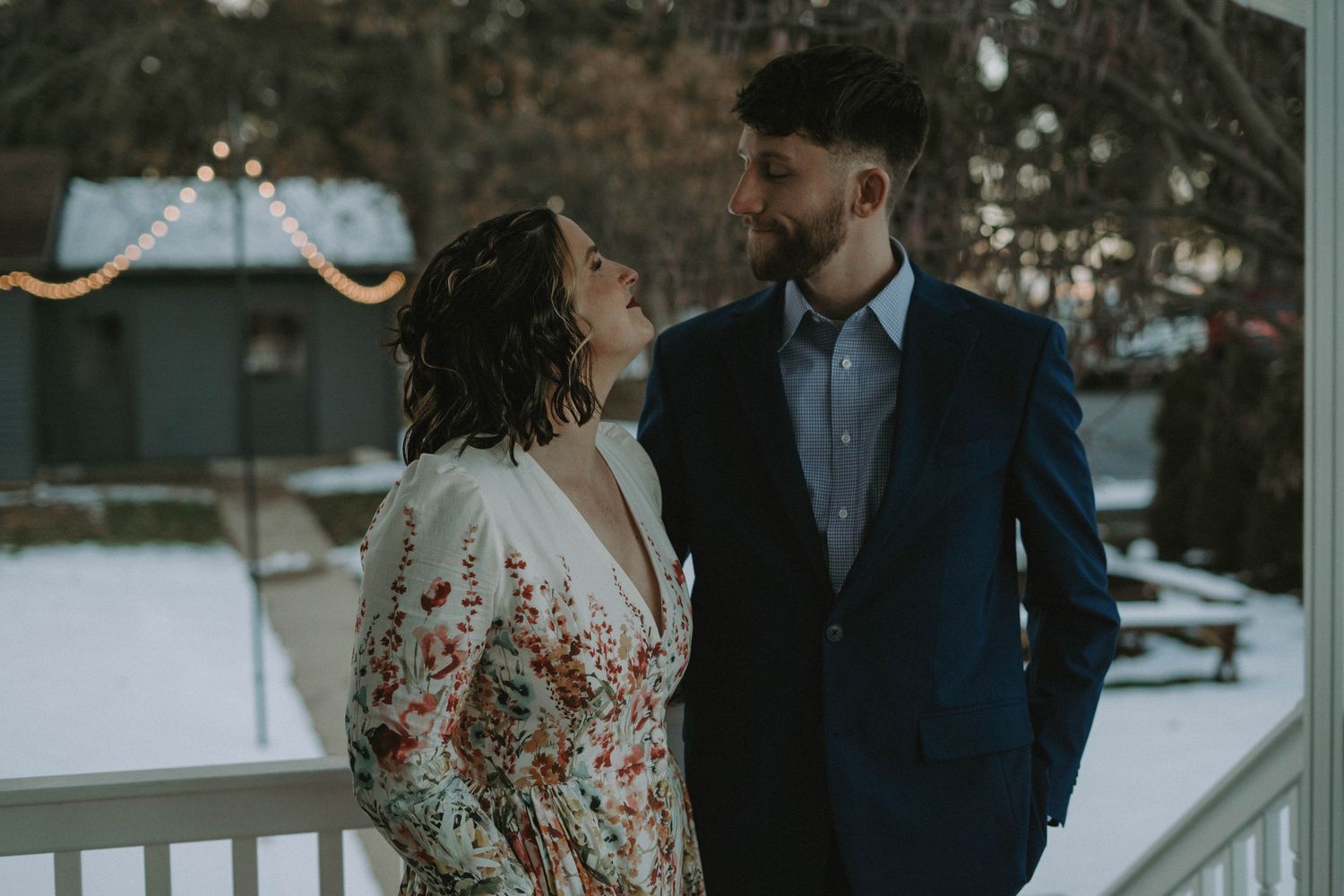 A couple in formal wear share an intimate moment on a snow-covered porch.