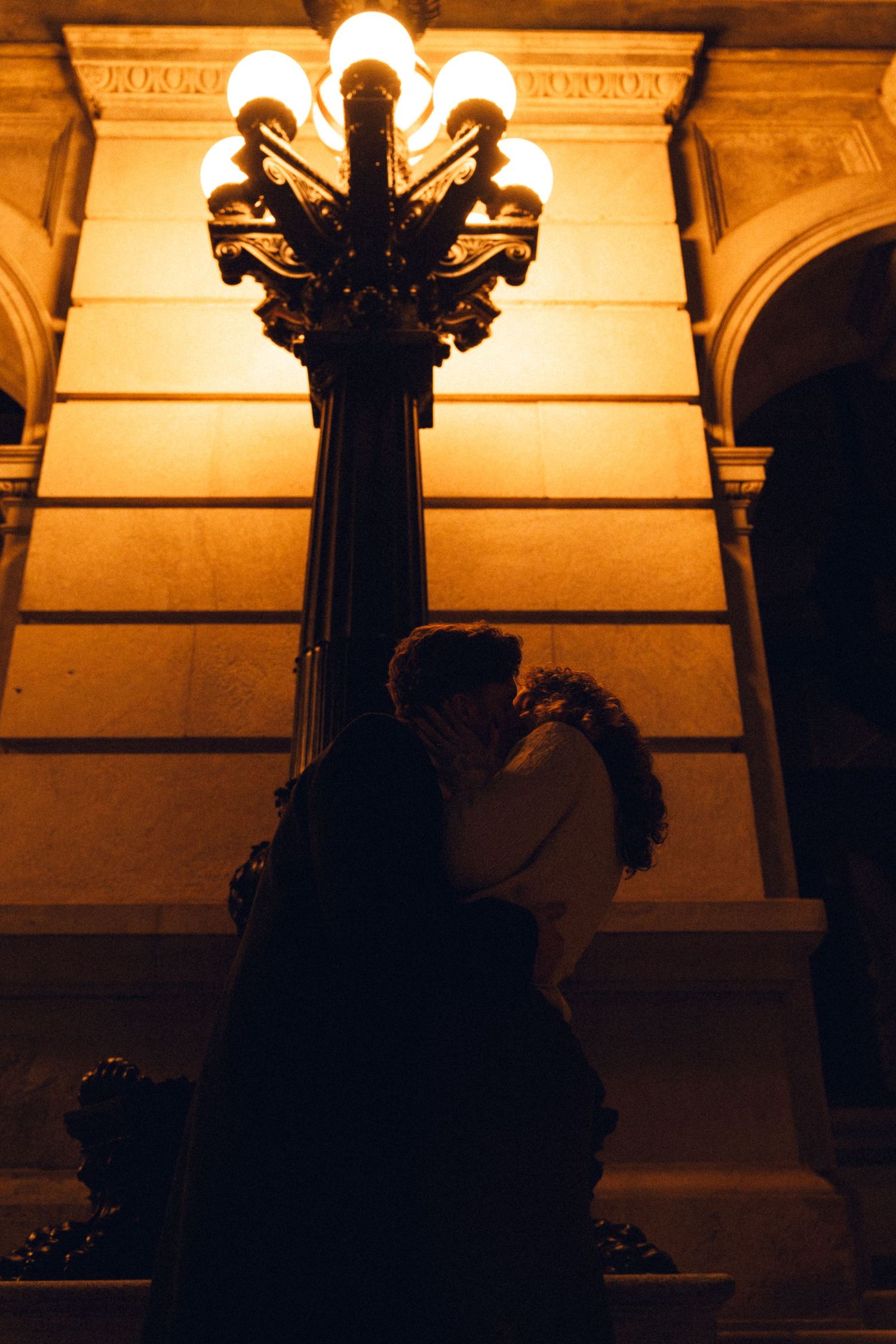 Ornate street lamps illuminate classical architecture columns at dusk creating a warm amber glow against arches.
