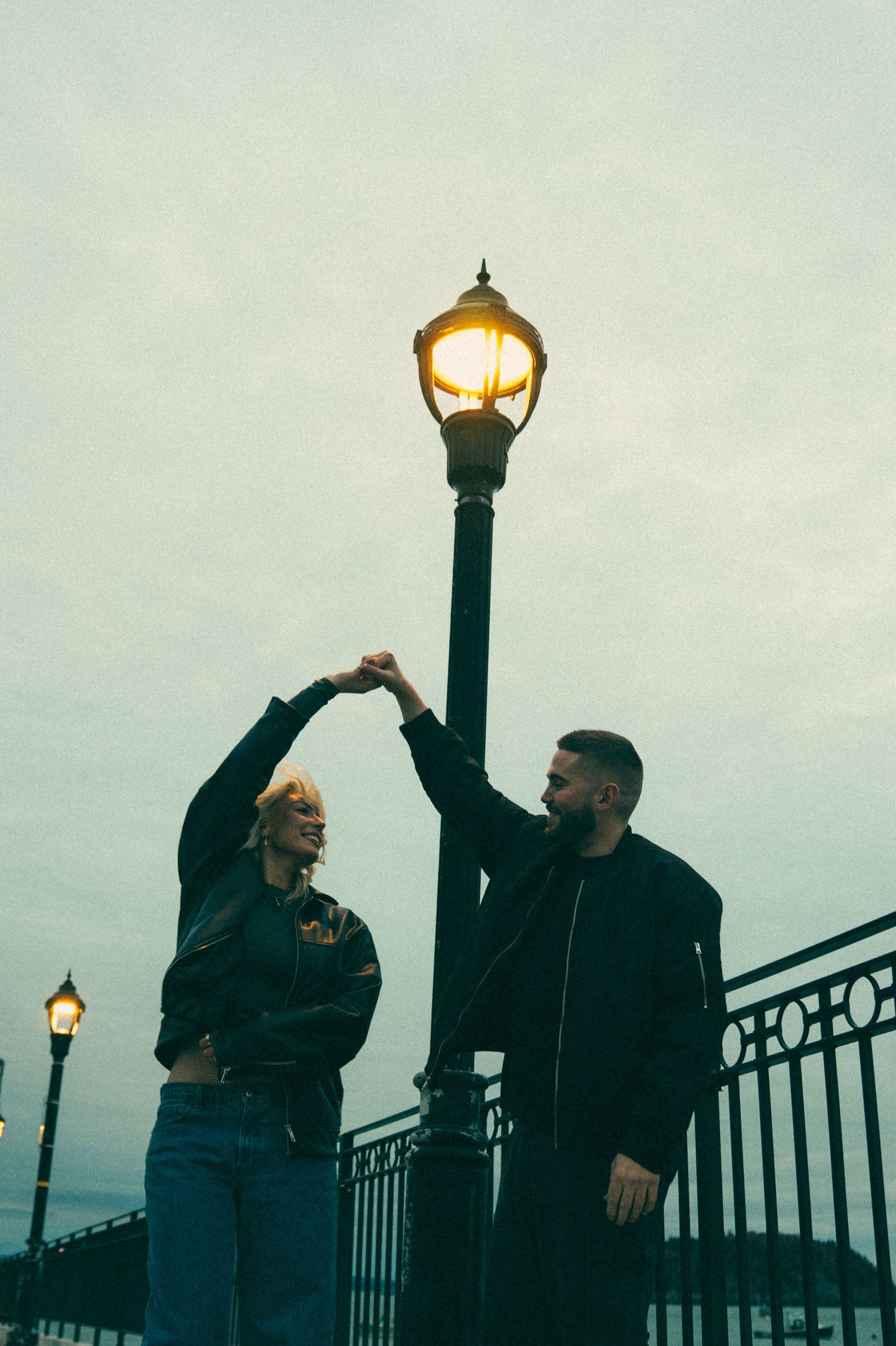 Two people in dark clothing pose against a railing with glowing street lamps at dusk.Couple in Bar Harbor Maine