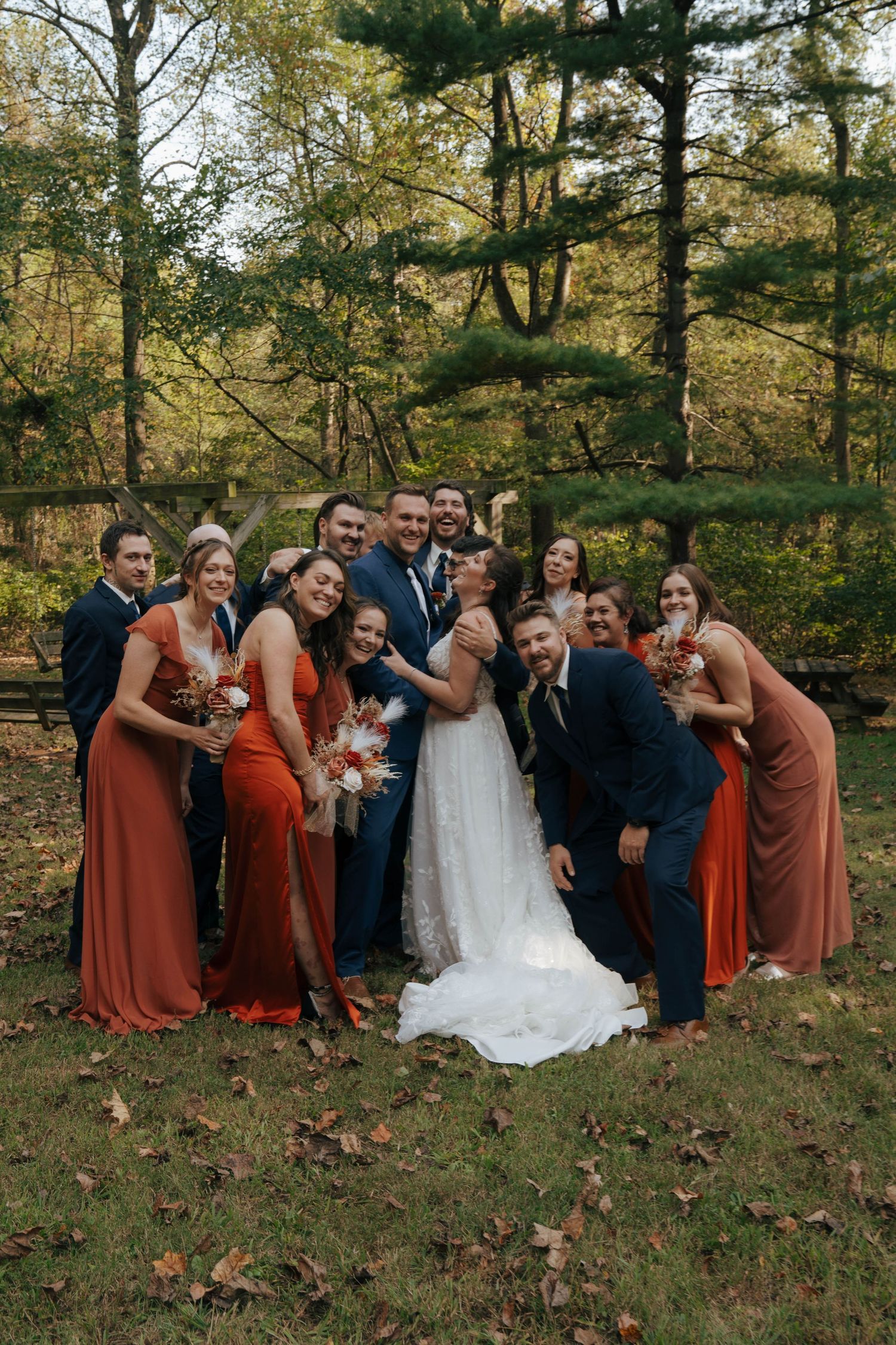 Wedding party in rust-colored dresses and navy suits pose together in autumn woodland setting.