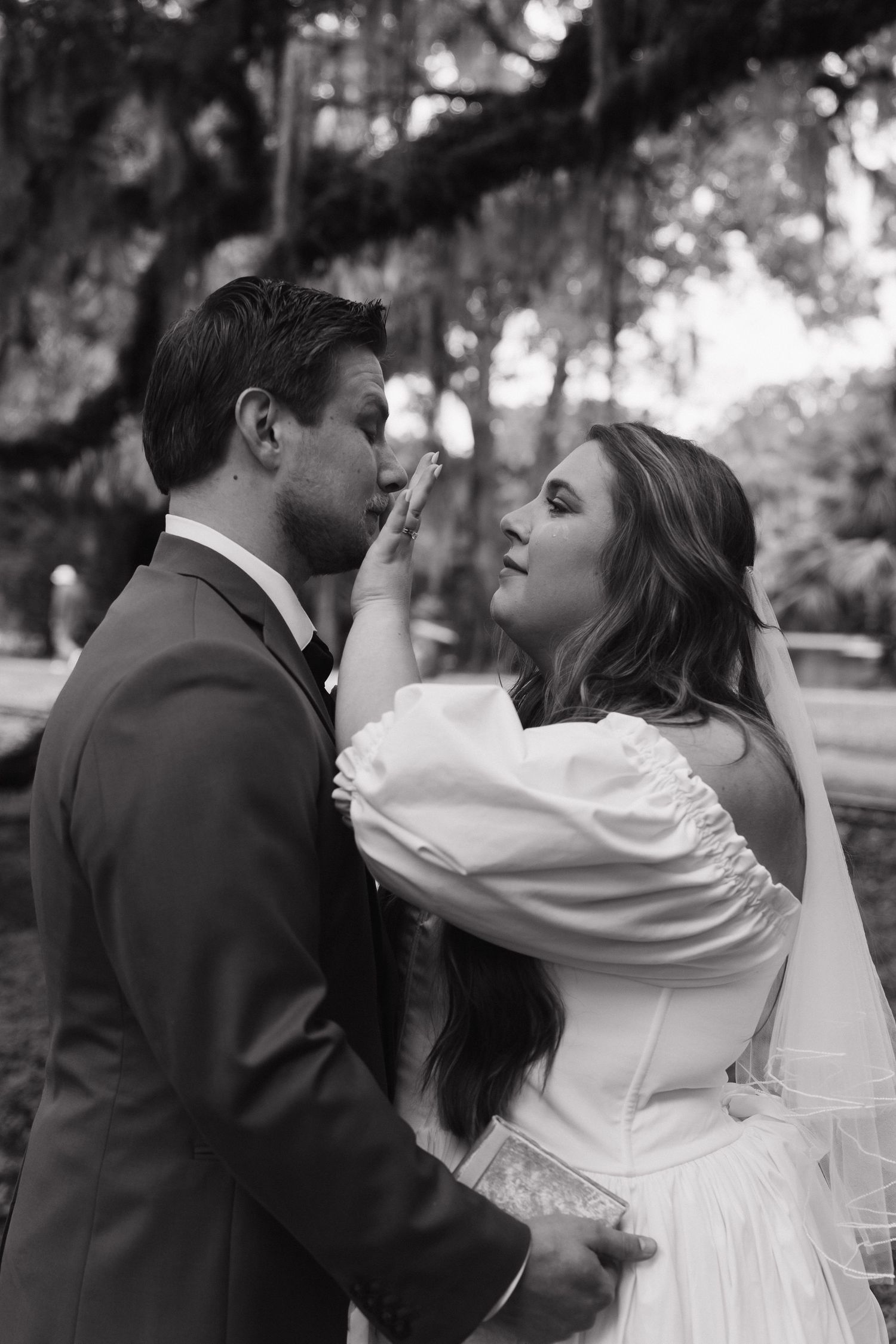 A romantic black and white wedding portrait of a couple in an intimate moment beneath trees with hanging moss.