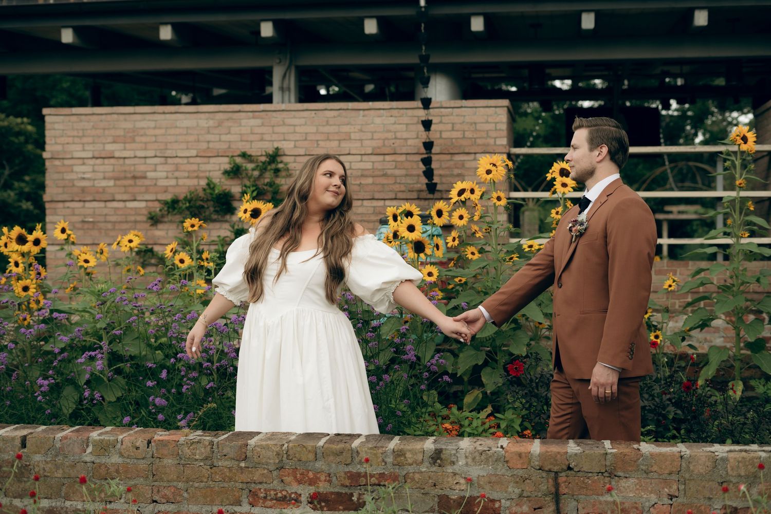 A couple in wedding attire holds hands in a garden with blooming sunflowers and brick walls.