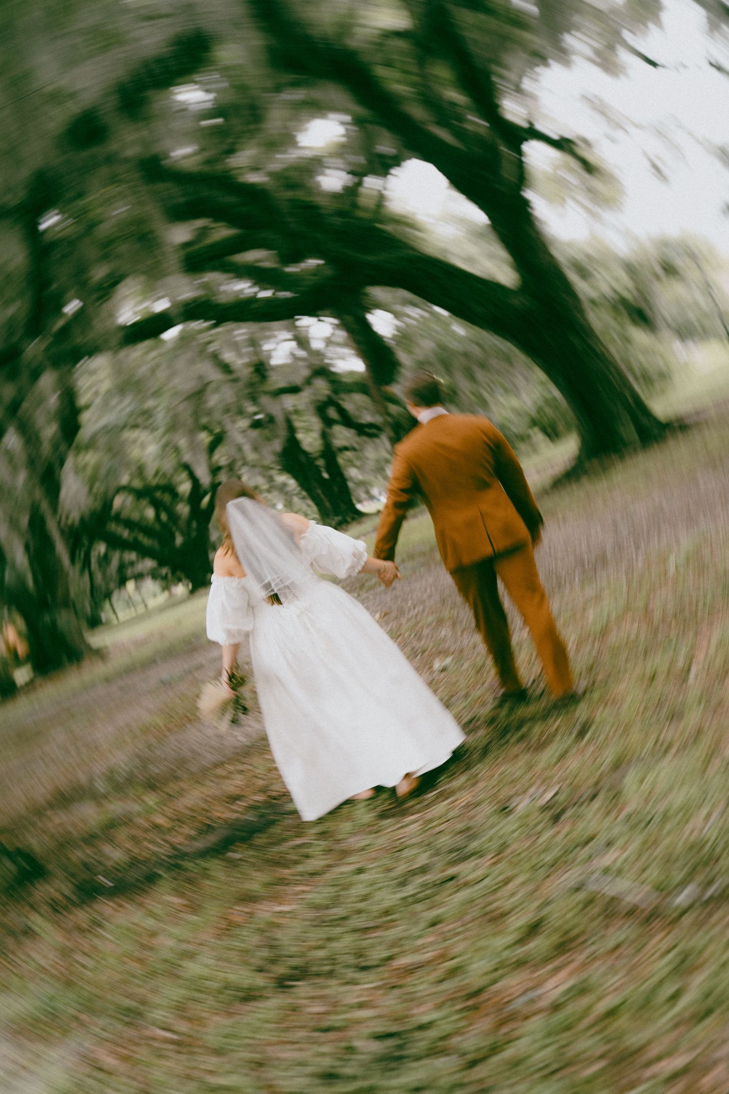 A couple in formal attire walks down a path beneath twisting tree branches in an artistic blur motion effect.