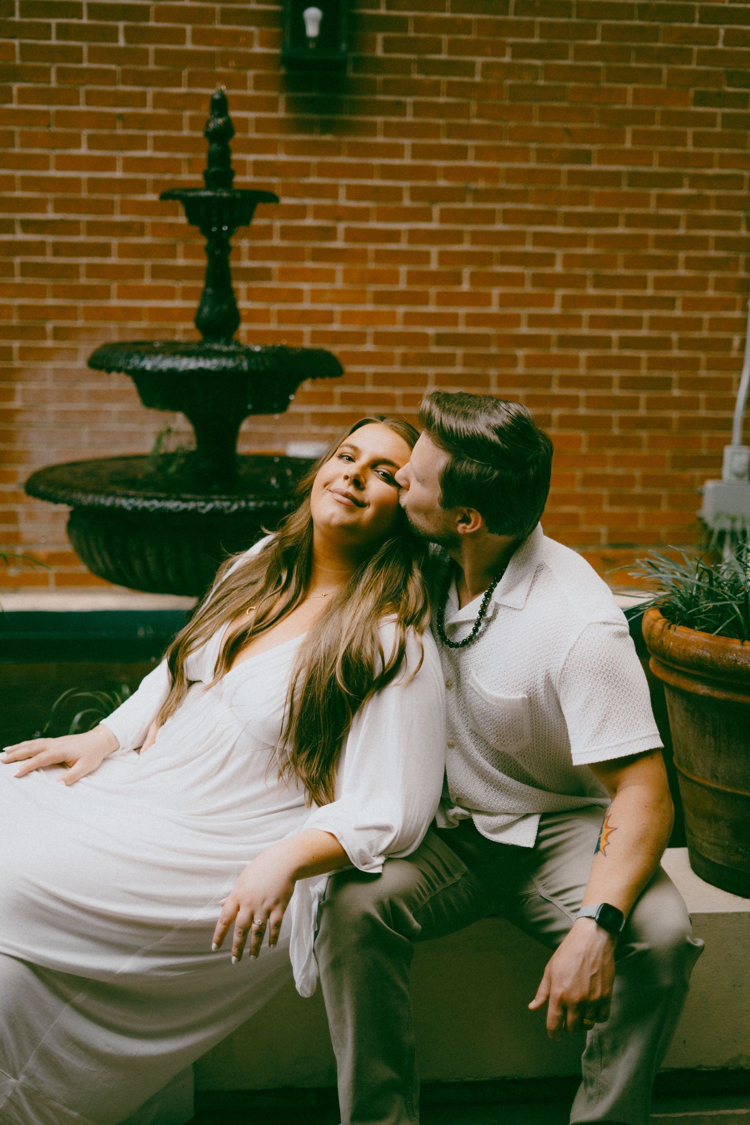 Maternity couple sharing tender moment on bench by vintage fountain and brick wall.