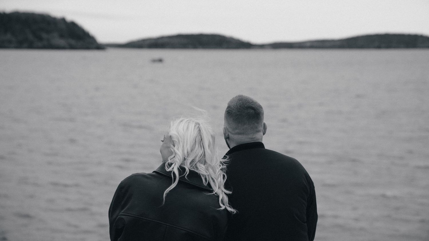 Elderly couple sitting together gazing at a calm sea in a black and white photo, with a small boat in the distance.