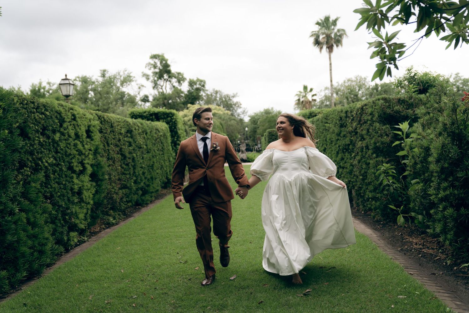 A couple runs joyfully down a garden path lined with tall green hedges as the bride's white dress flows behind them.
