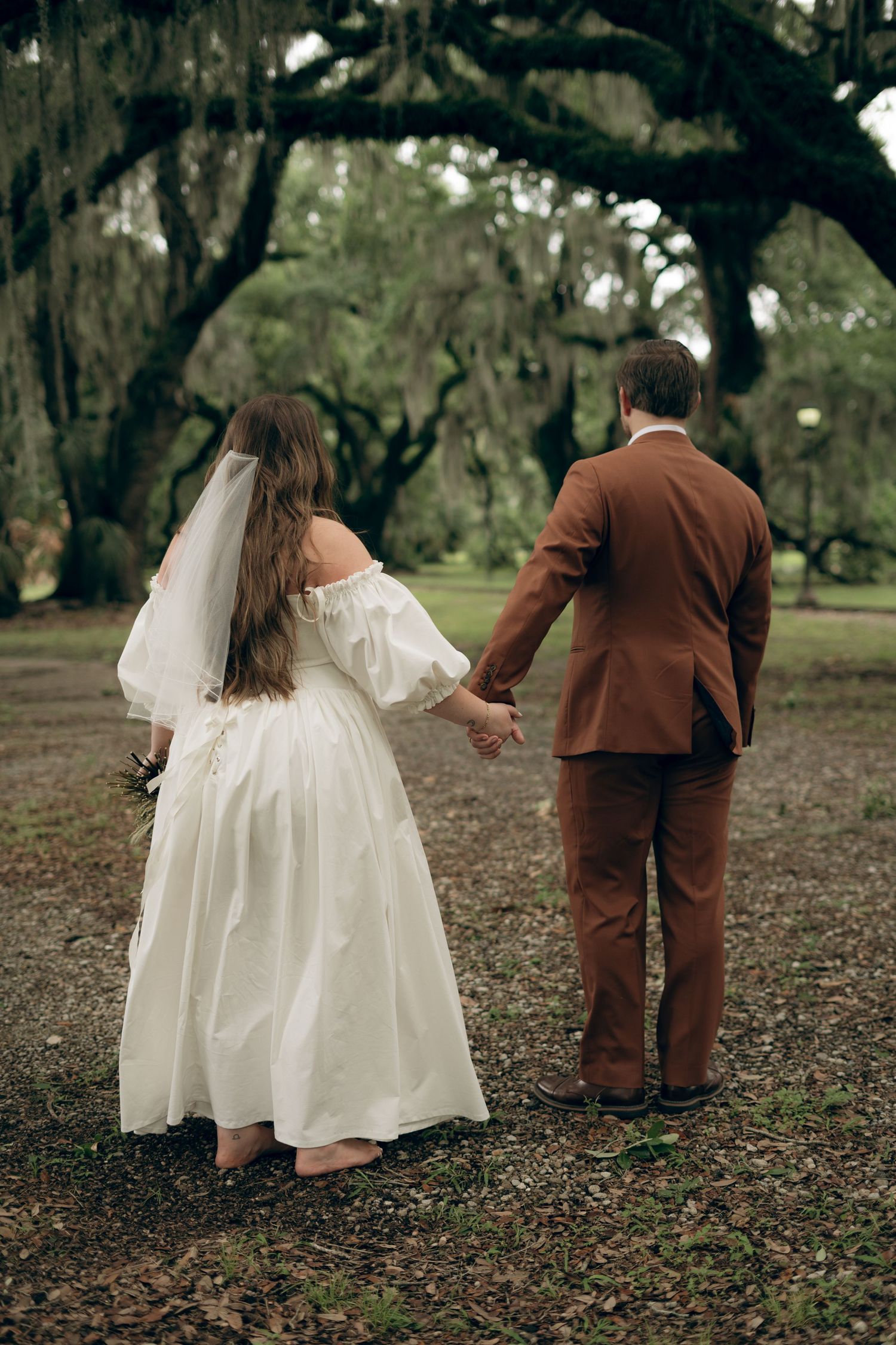 A bride in a white off-shoulder gown holds hands with her groom in a brown suit under draped oak trees.