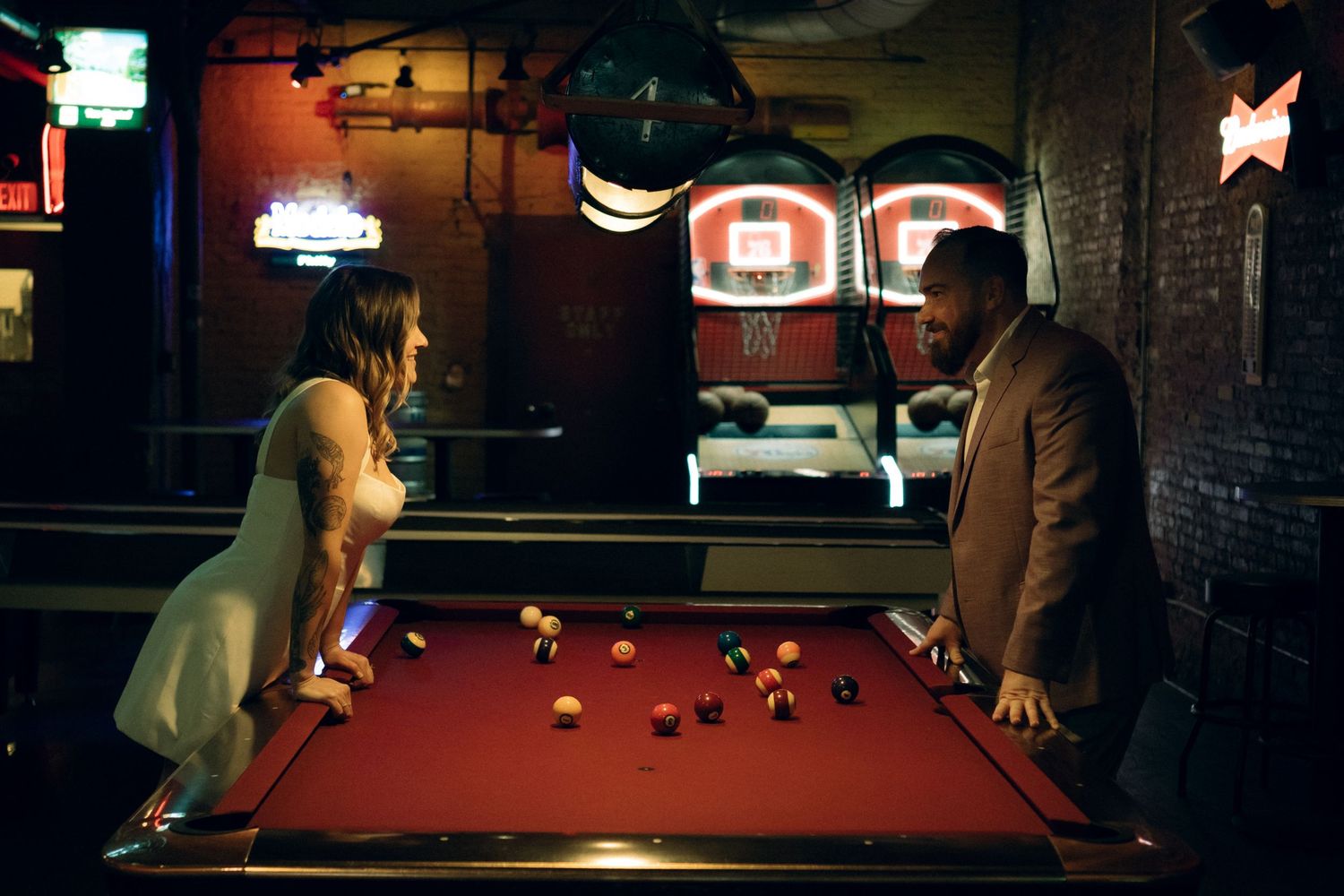 Two people playing pool in a dimly lit bar with neon signs on the brick wall in the background.