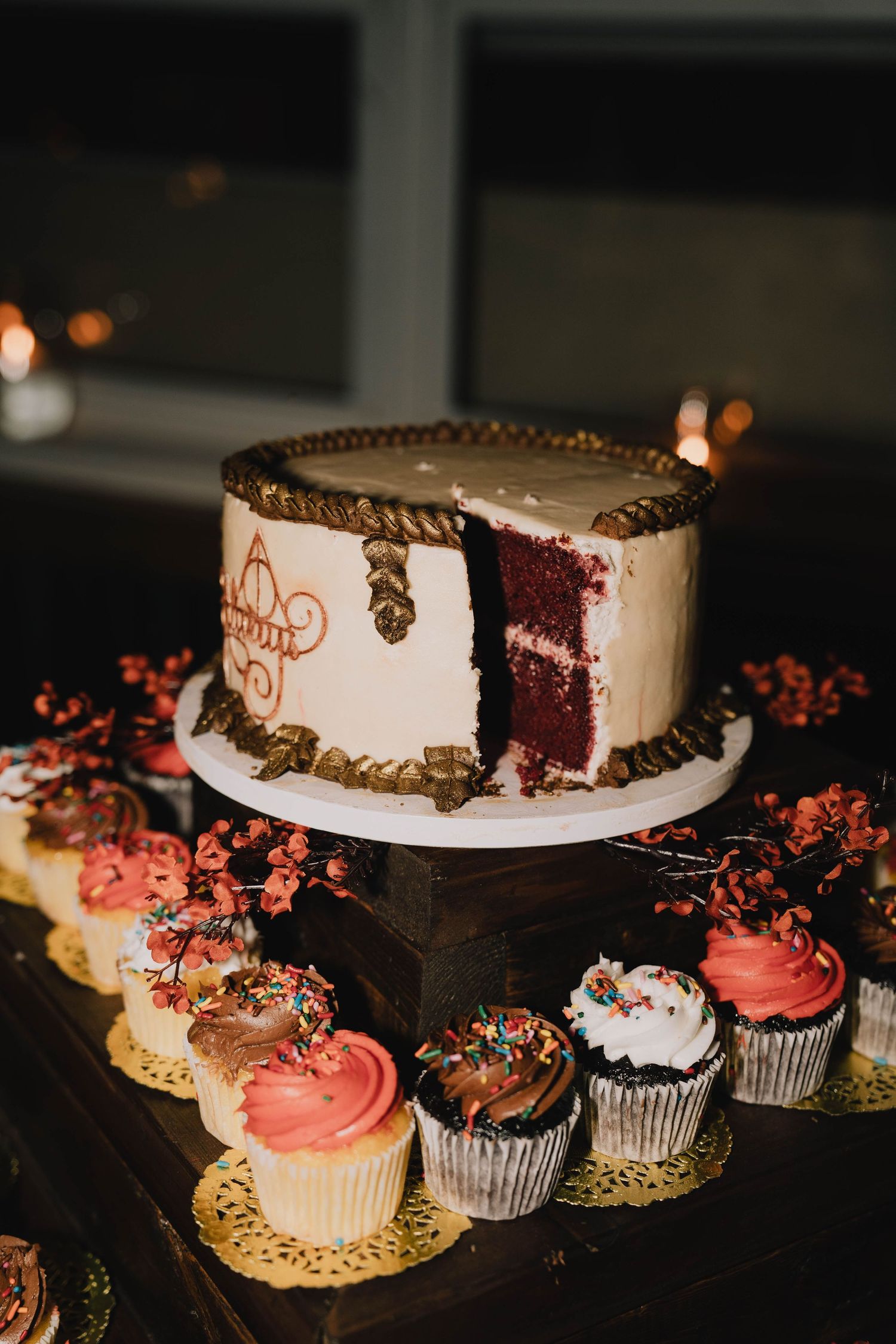 Elegant white and gold wedding cake with red velvet interior alongside decorated cupcakes on a dark display.