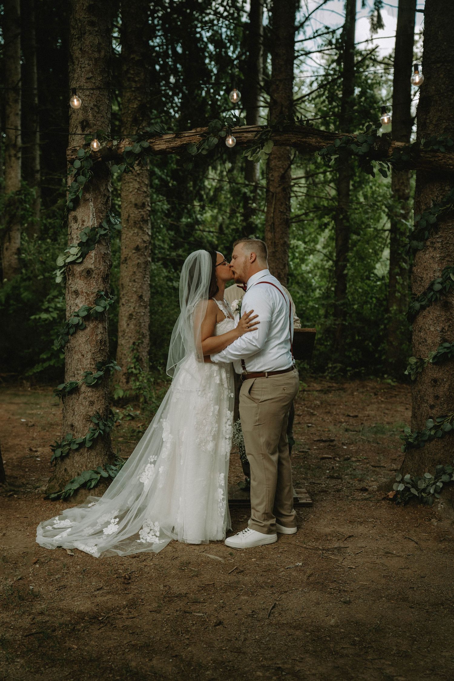 A romantic kiss under string lights in a magical forest wedding setting.
