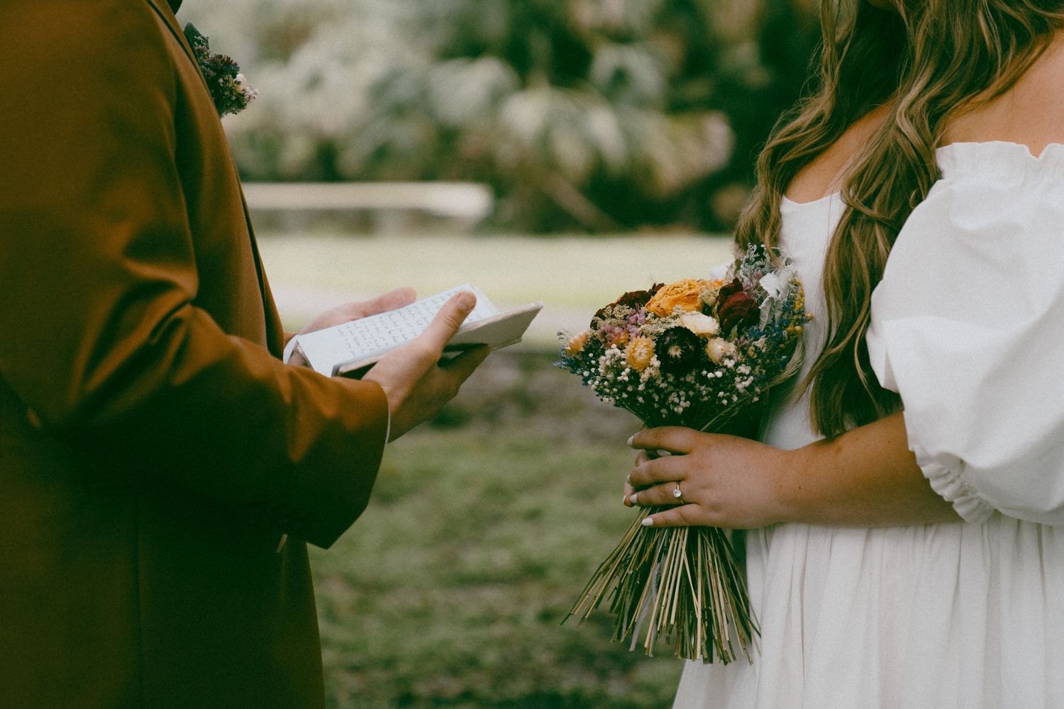 A wedding ceremony outdoors with hands holding vows and a rustic wildflower bouquet in white and green tones.