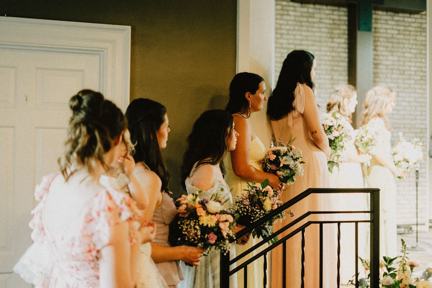 Bridesmaids holding elegant floral bouquets stand in line on stairs at a vintage-style wedding venue.
