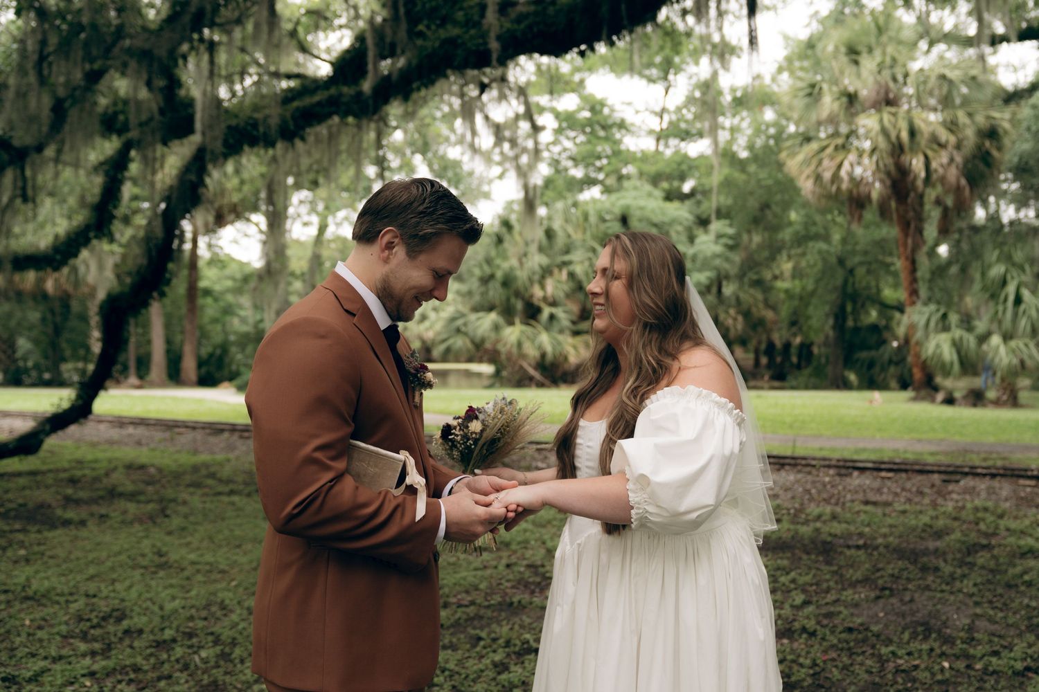 Two people in romantic dress stand together outdoors in a garden setting with Spanish moss and palm trees in the background.