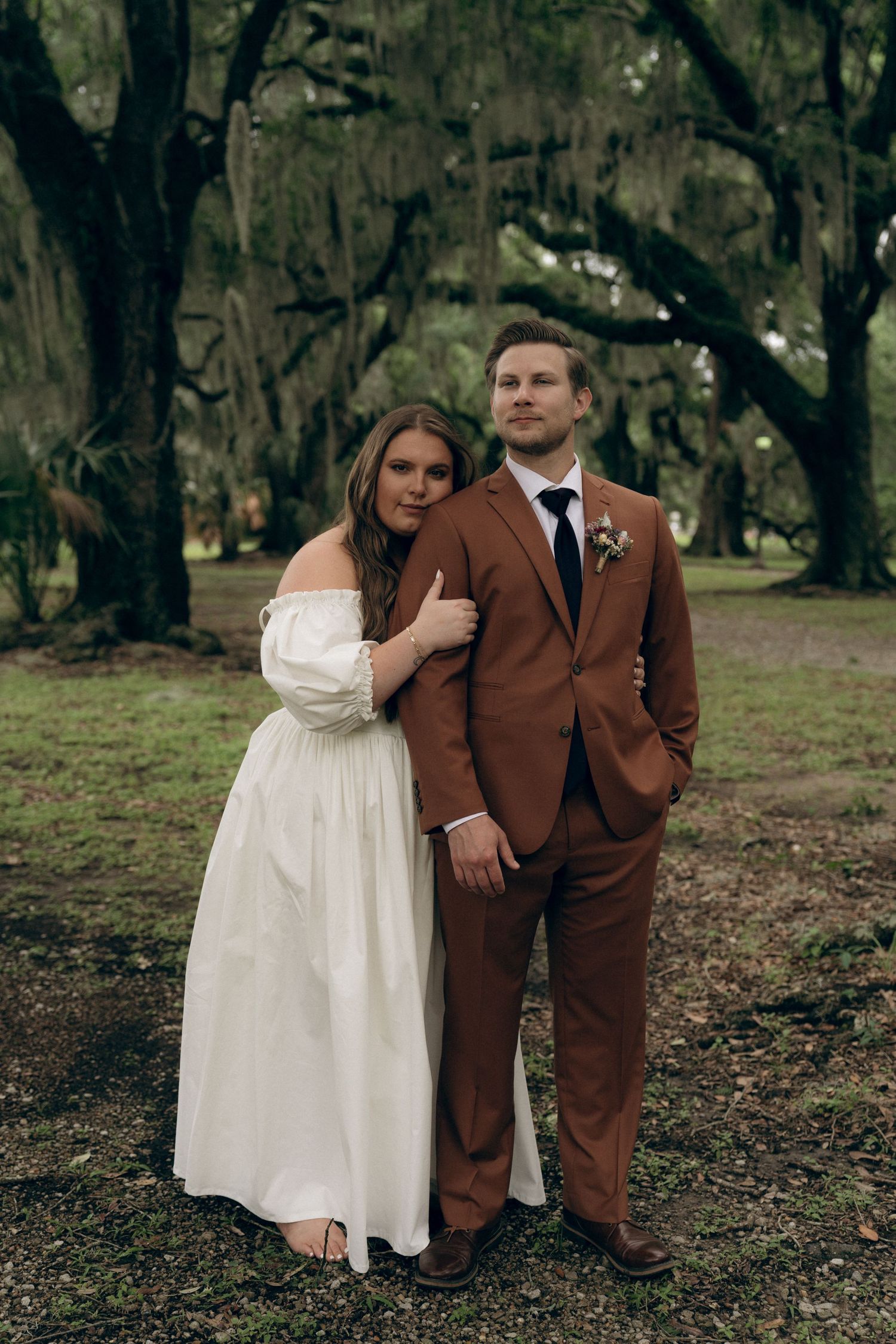 Couple in wedding attire poses under moss-draped oak trees, with bride in white dress and groom in brown suit.
