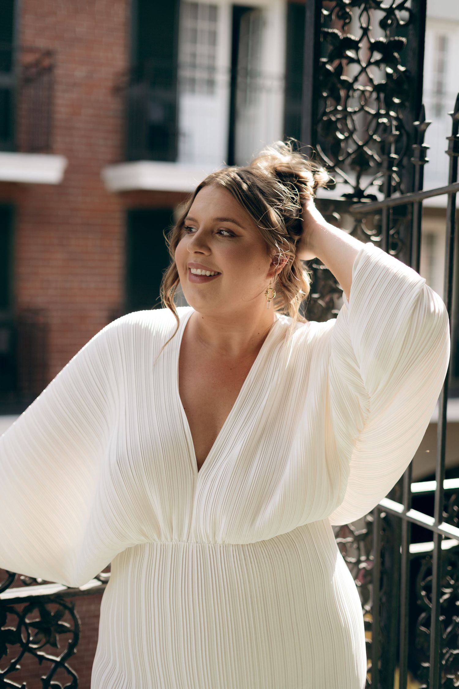 Person in white pleated dress with bell sleeves poses joyfully against ornate iron gating and brick building exterior.
