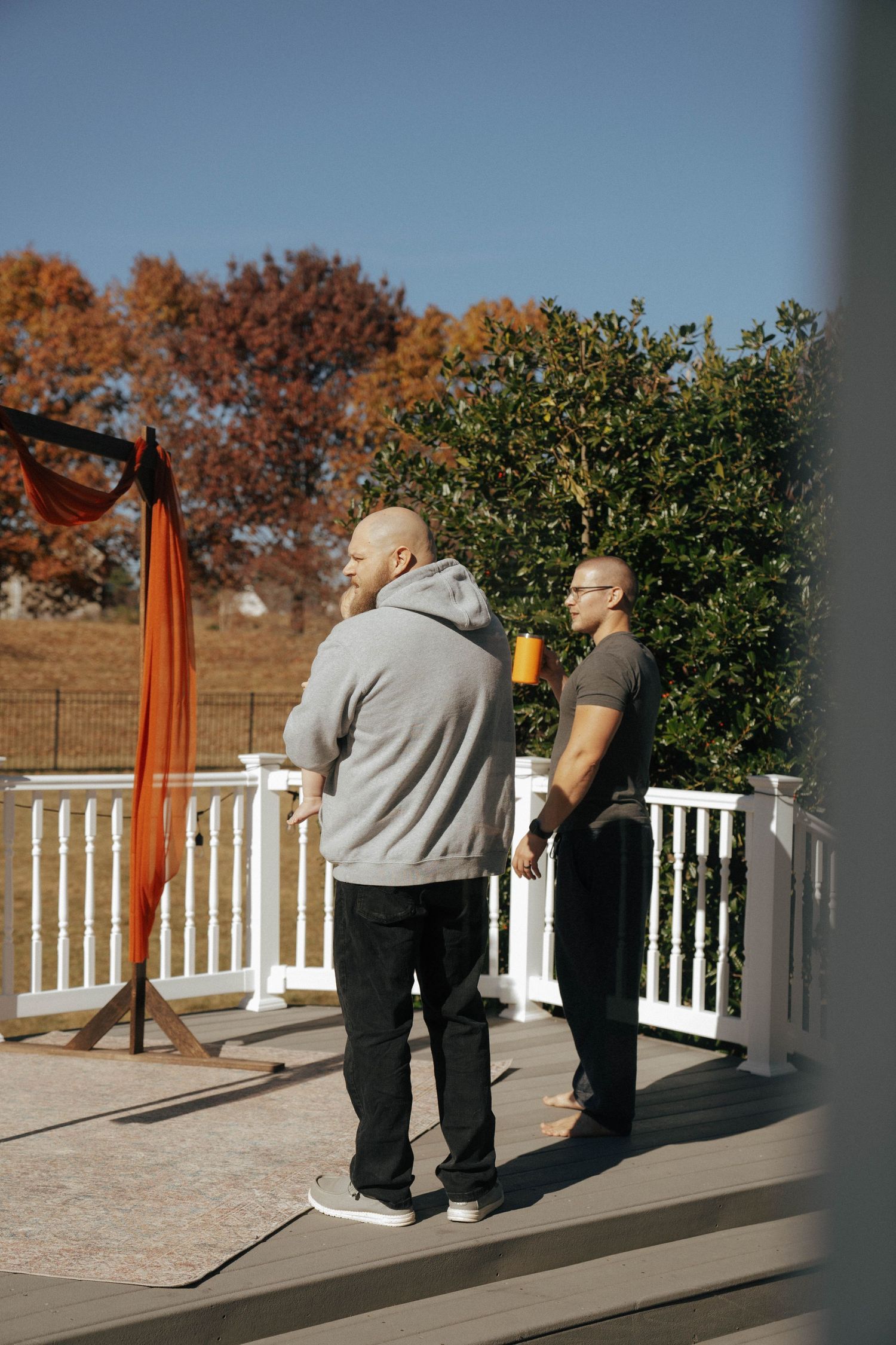 Two people in casual attire stand near an orange-draped wedding arch on a white deck.