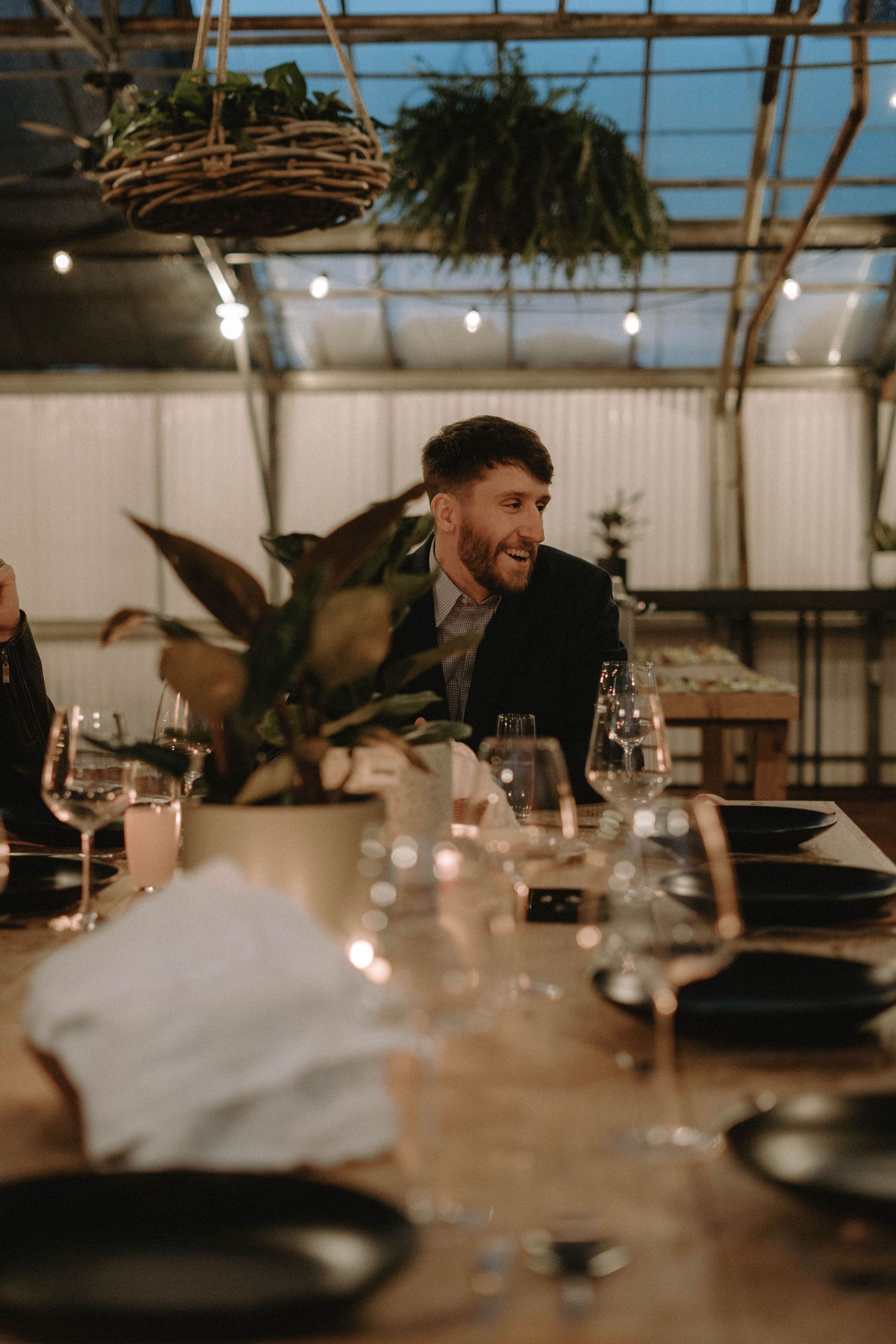 Guests enjoying laughter during an elegant dinner party under hanging plants and ambient lighting.