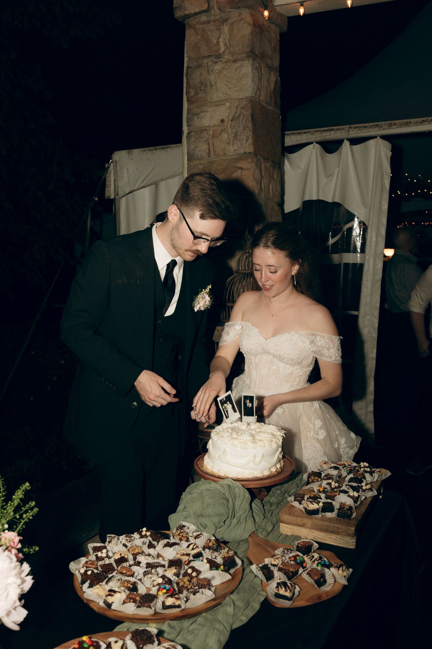 A couple in wedding attire cut their white wedding cake together at their reception dinner while surrounded by desserts.