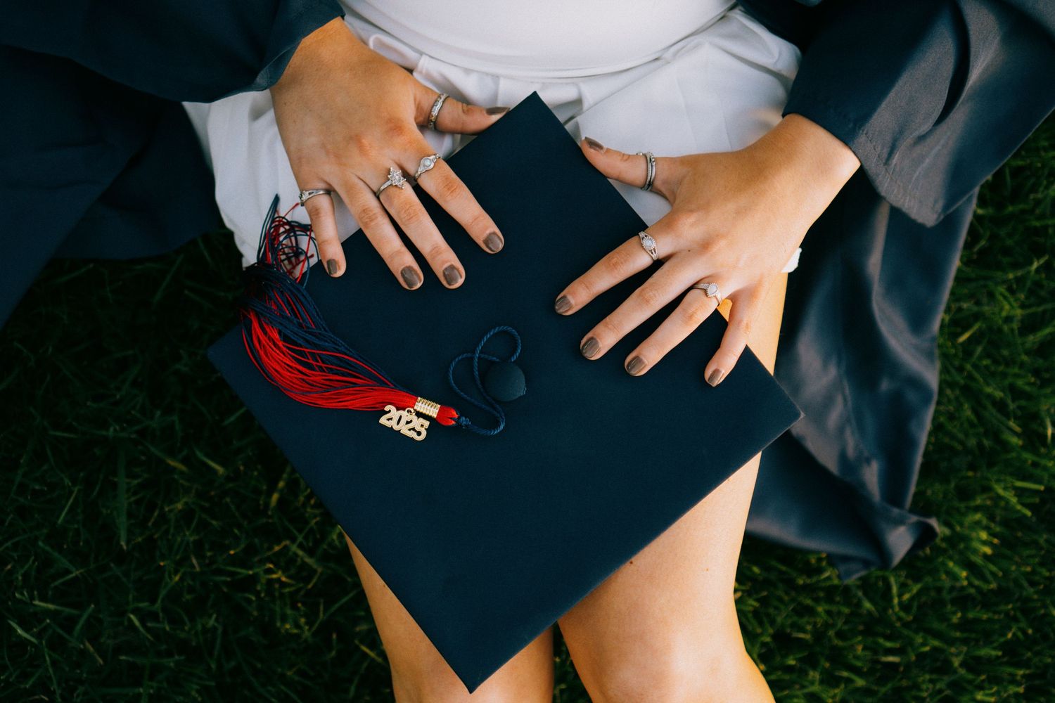 Close-up view of hands touching a navy graduation cap with red tassel while sitting on grass.