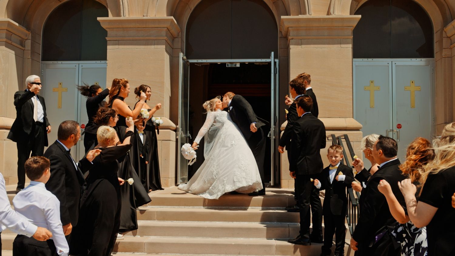 Bride and groom share a kiss on church steps surrounded by wedding party in elegant black attire at outdoor ceremony.