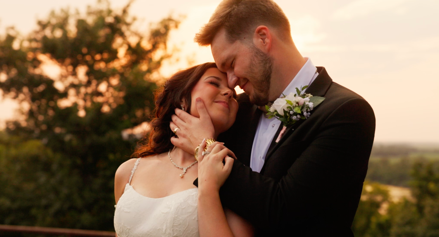 Romantic wedding couple embracing at sunset, bride in white gown and groom in black tuxedo with boutonniere, outdoors.