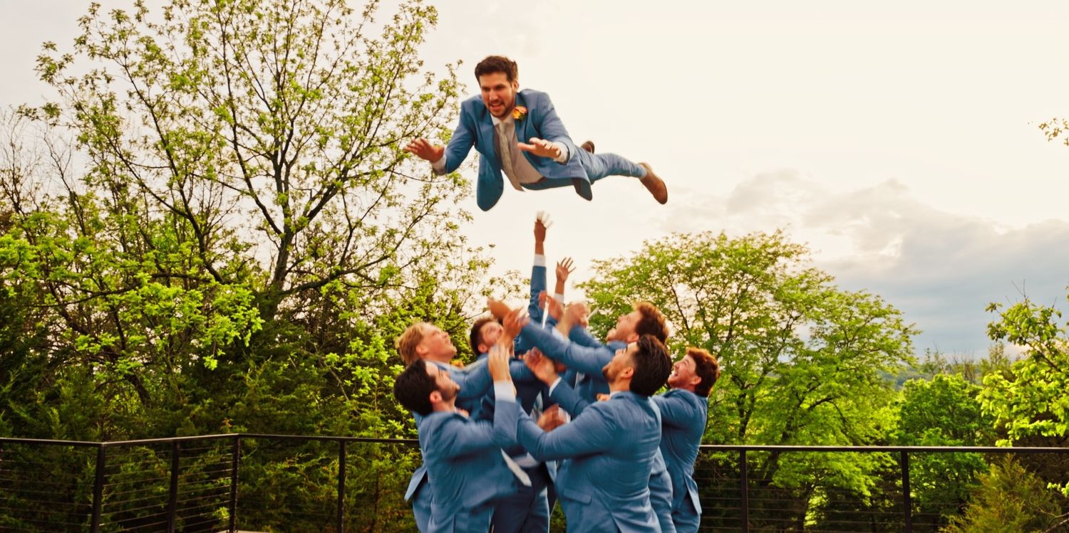 Groomsmen tossing groom in the air outdoors, wearing matching blue suits, celebrating at a wedding.