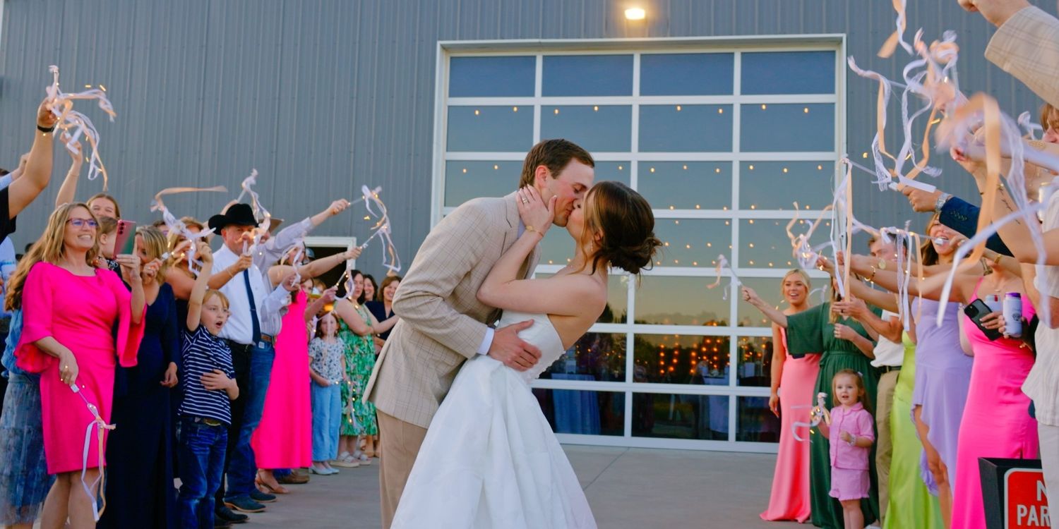 Bride and groom share a romantic kiss during wedding send-off with guests waving ribbon wands outside a modern venue.