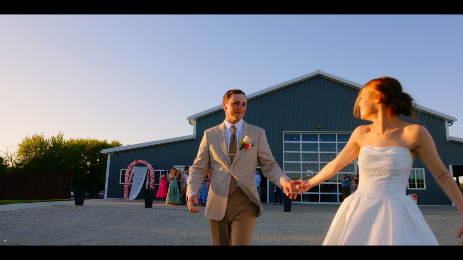 Bride and groom holding hands walking toward camera at outdoor wedding venue during golden hour sunset.