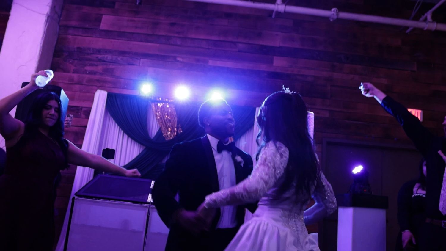 Bride and groom share first dance at elegant wedding reception with purple stage lighting and wood accents.
