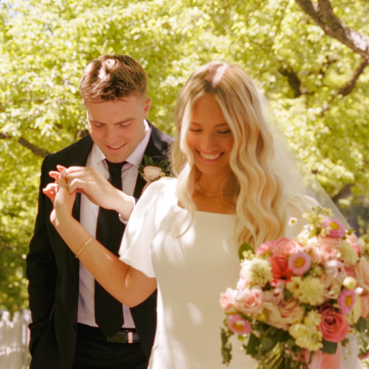 A bride in a white gown holds pink florals beside a groom in a black suit, smiling outdoors under green trees.