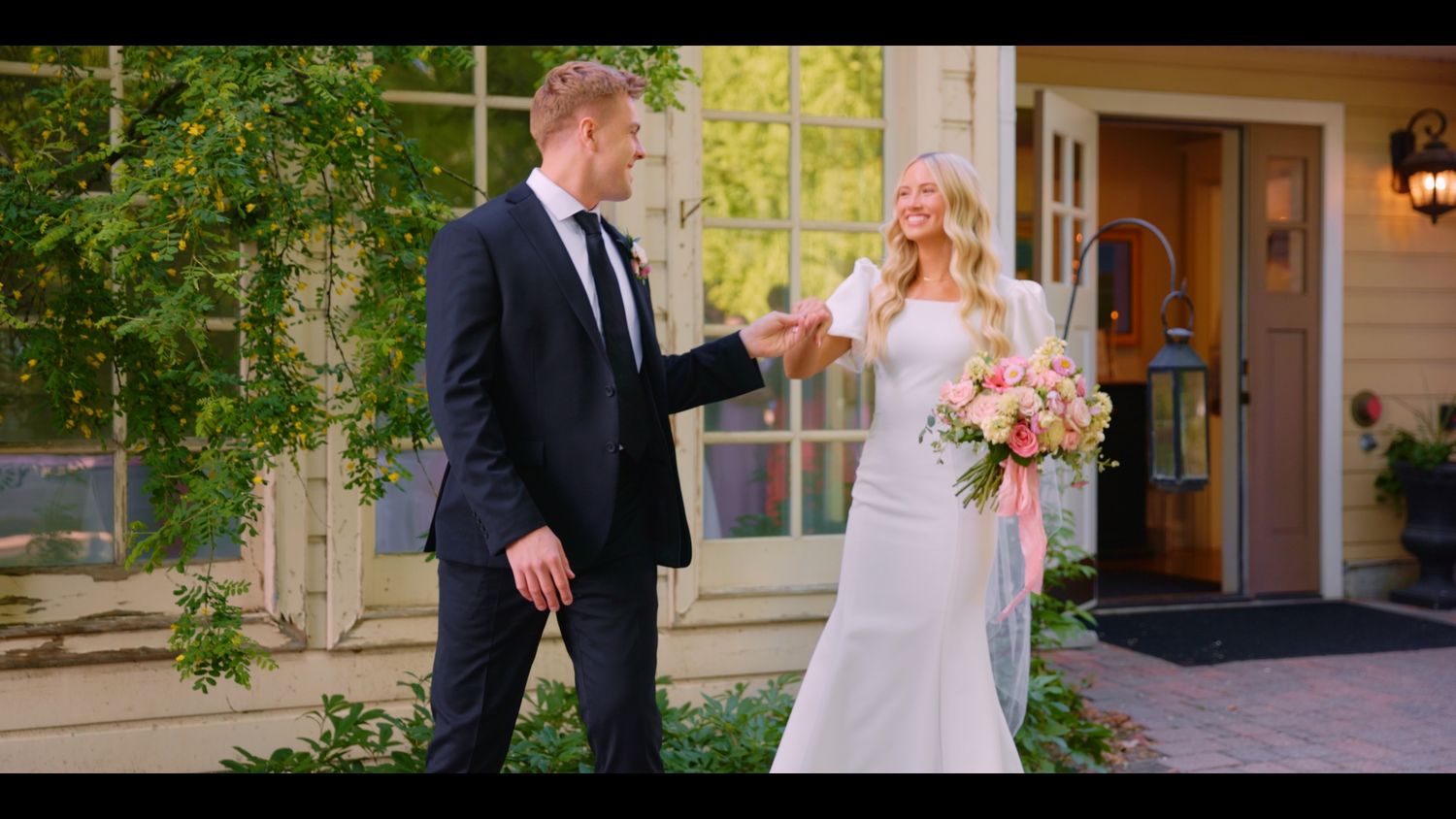 Bride in white gown holding pink bouquet meets groom in navy suit outside elegant venue.