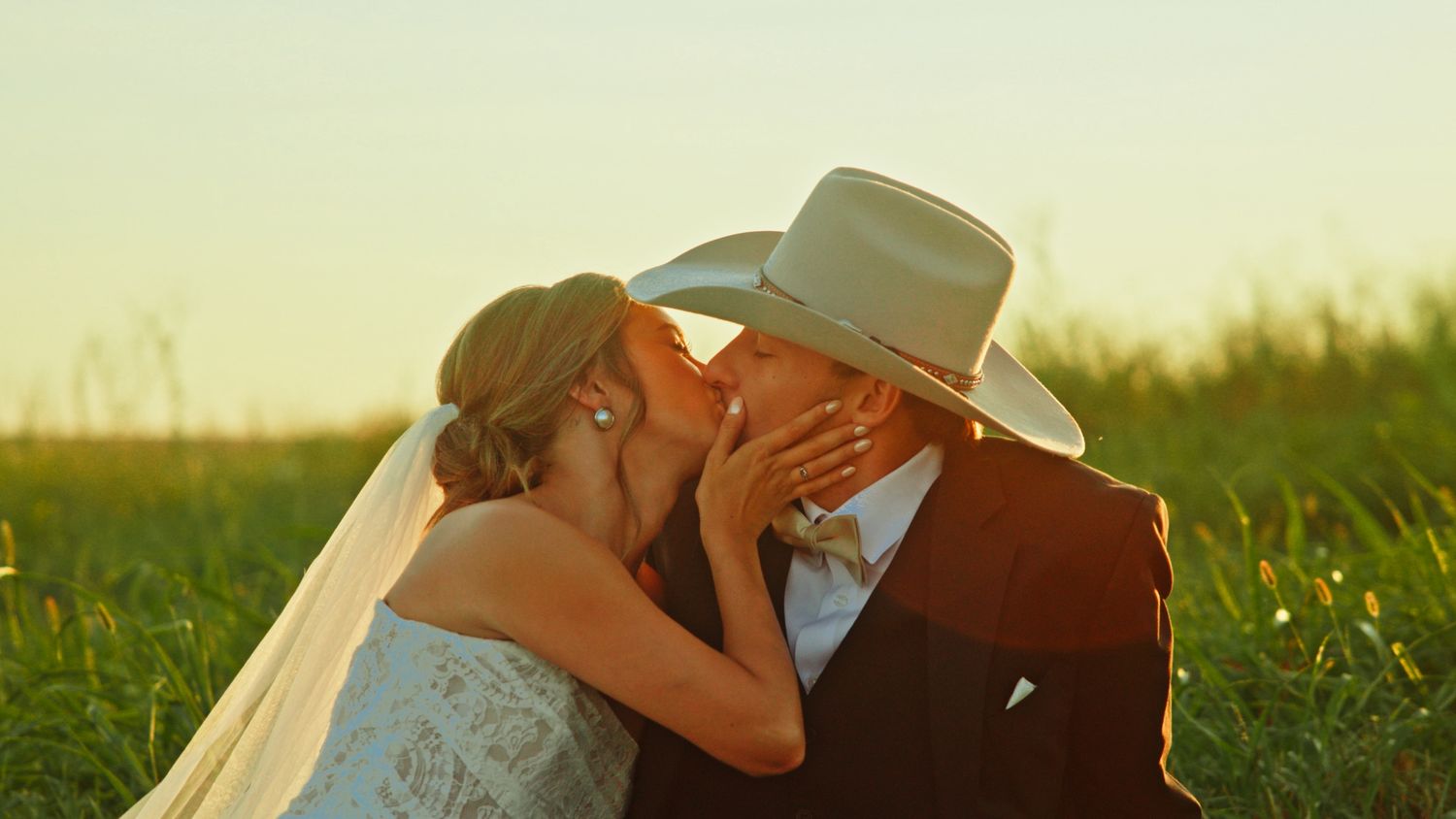 A bride and groom share a romantic kiss in a sunlit field, the groom wearing a cowboy hat and brown suit.